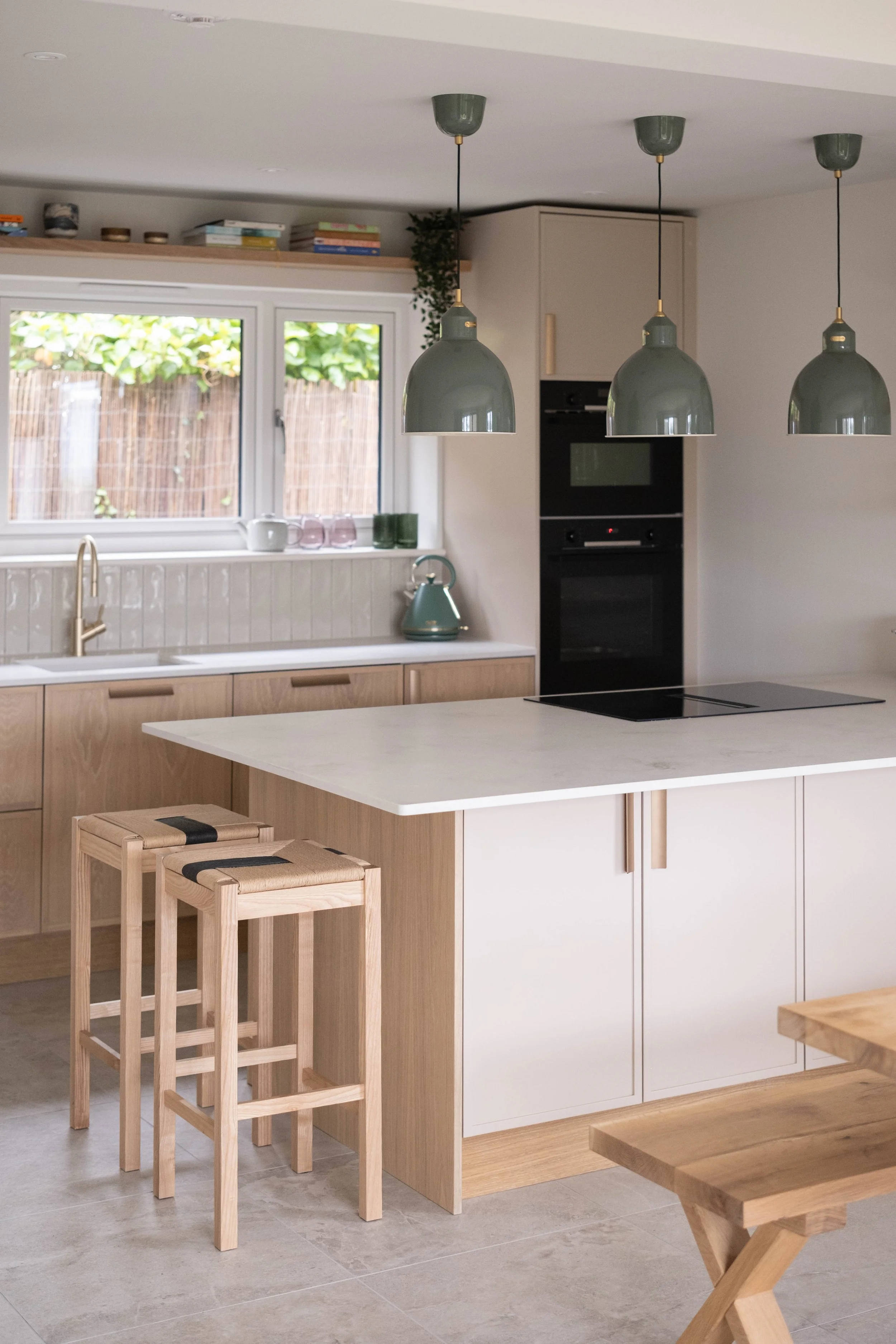 Modern kitchen with pendant lights, a window with a view of a wooden fence and greenery, a light wood kitchen island with stools, and a black oven built into cabinetry.