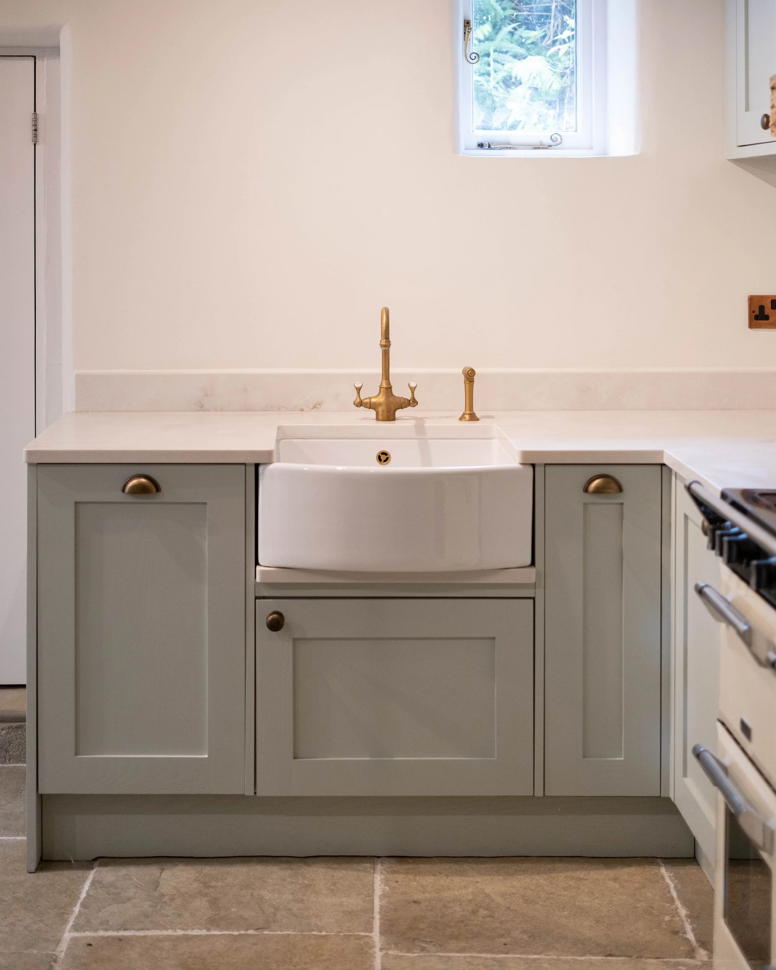 A kitchen sink area with a white farmhouse sink, a brass faucet, and a light gray cabinet below. There's a small window above the sink allowing natural light, with a view of greenery outside. The floor is tiled with large beige tiles.