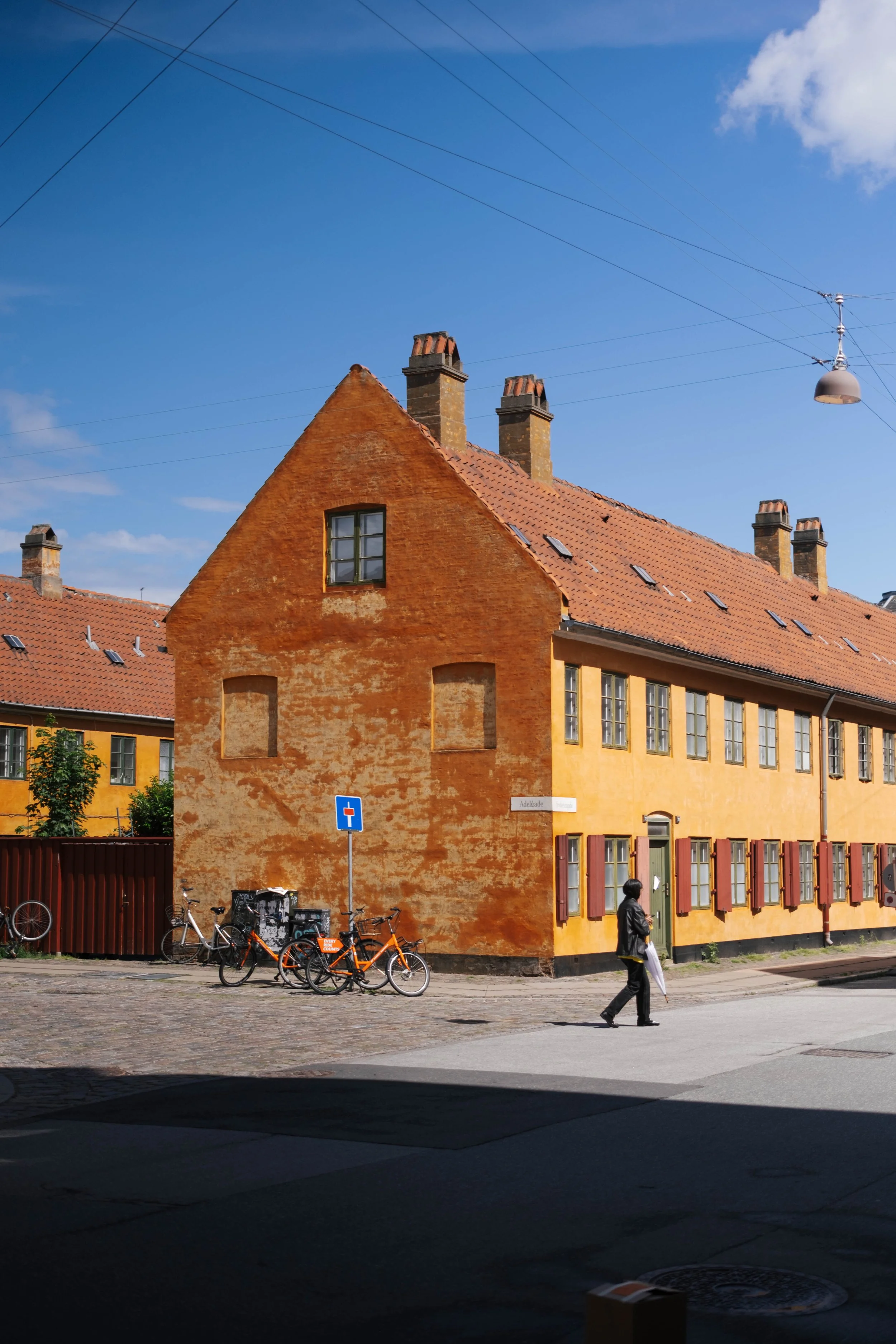 A street scene featuring a large orange brick building with multiple chimneys, red-tiled roof, and several windows. Bicycles are parked in front, and a person is walking across the street. Overhead tram wires are visible against a bright blue sky wit