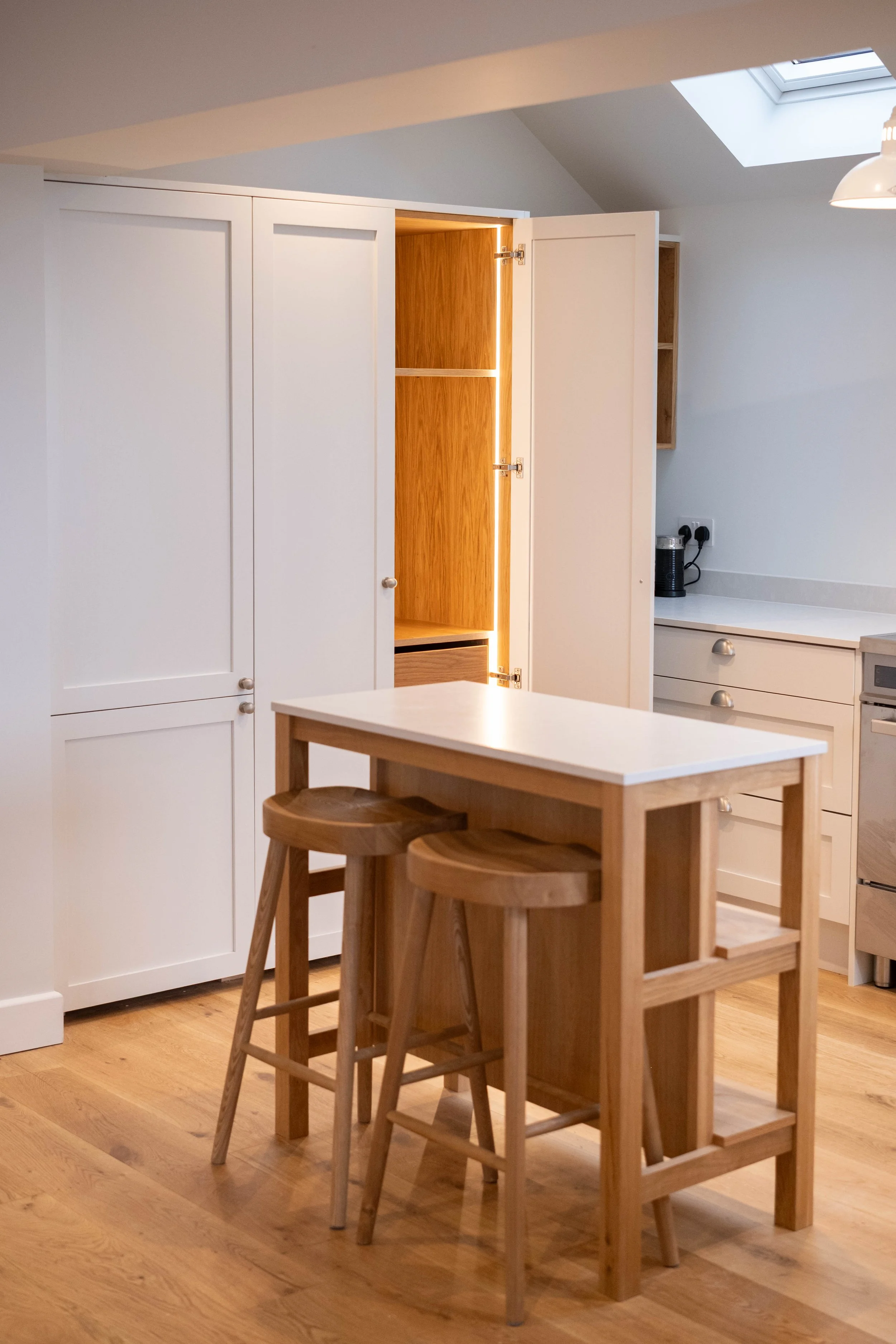 A cozy kitchen with white cabinets, a small wooden dining table, and three wooden stools. There is a skylight in the ceiling and a partially open cabinet with wooden interior, a countertop with a small appliance, and a plug socket on the wall.