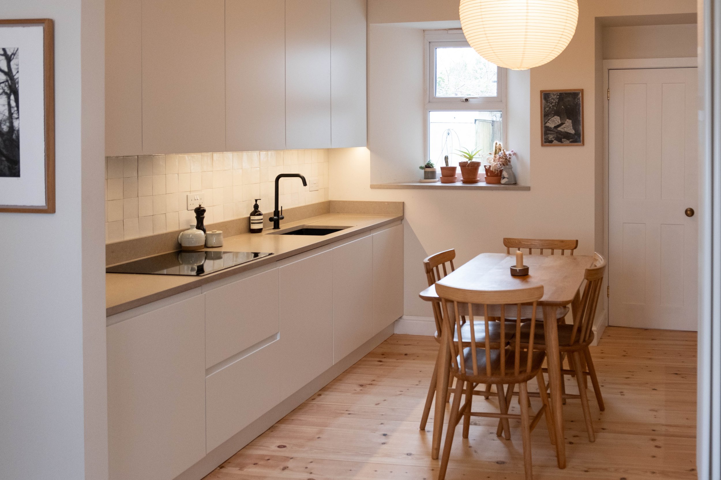 Modern kitchen with beige cabinets, a beige countertop, a black sink faucet, a small window with potted plants, a wooden dining table with four chairs, and a paper lantern overhead.