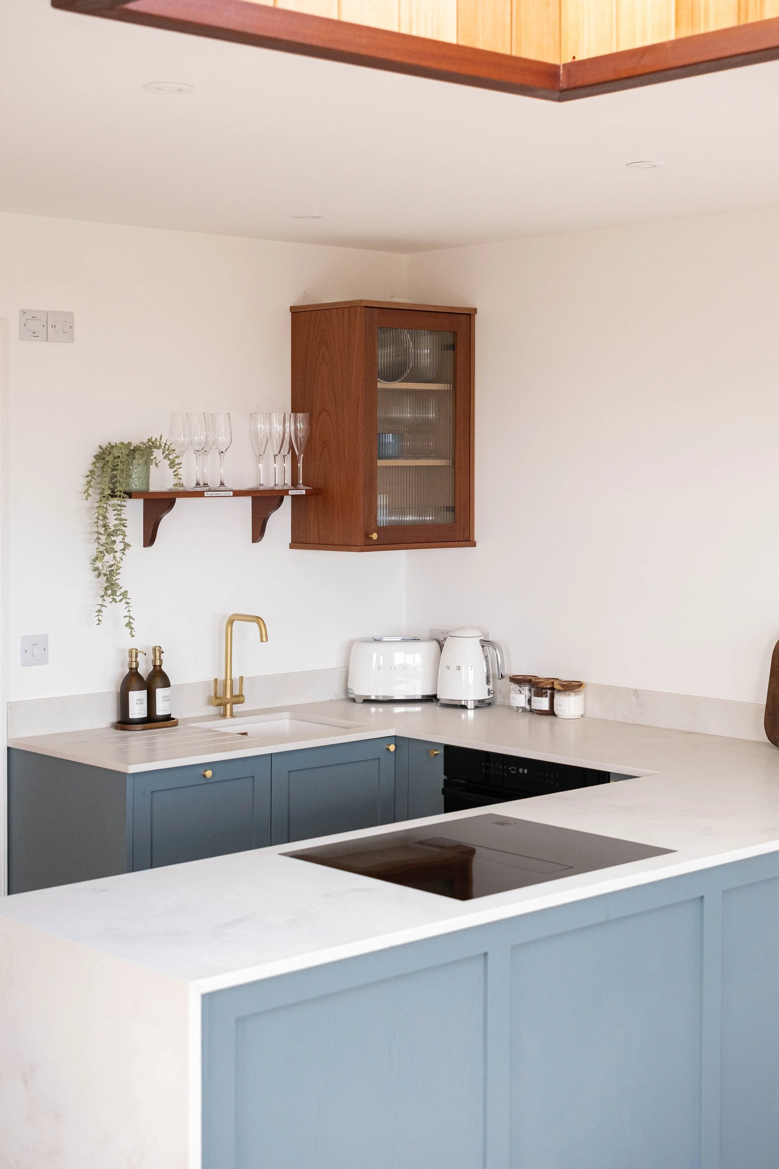 Modern kitchen with blue cabinetry, white countertops, brass faucet, wooden wall cabinet with glass door, floating shelf with glassware, potted plant, and kitchen appliances.