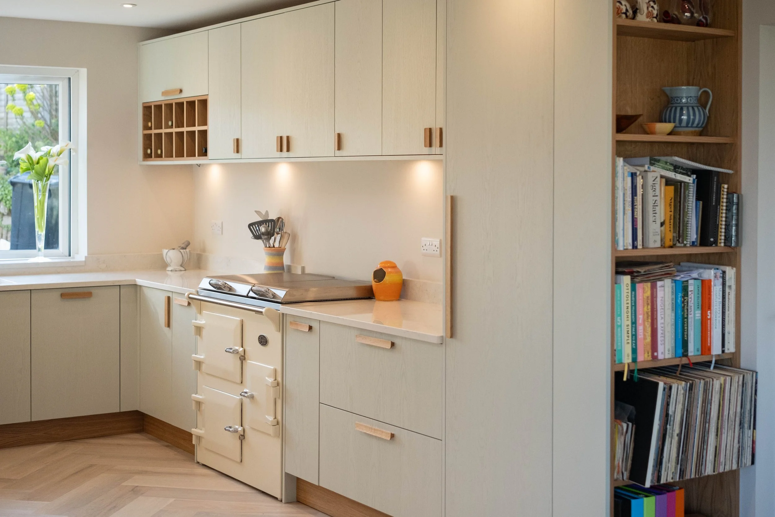 Modern kitchen with cream-colored cabinets, a window with flowers, a vintage stove, and a bookshelf filled with books and decorative items.