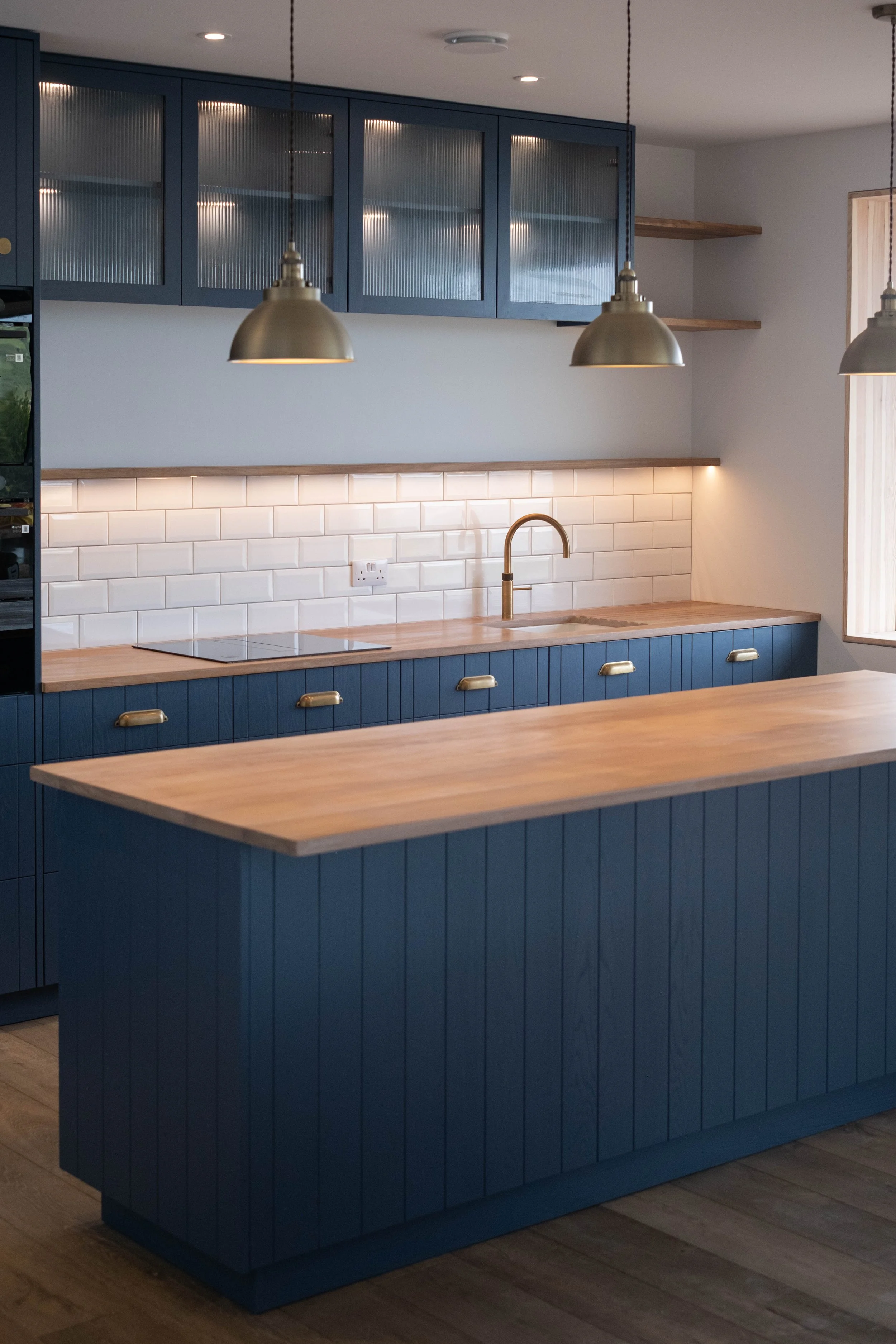 Modern kitchen with blue cabinetry, wooden countertops, white subway tile backsplash, and hanging pendant lights. There is a gold faucet and an electric cooktop.
