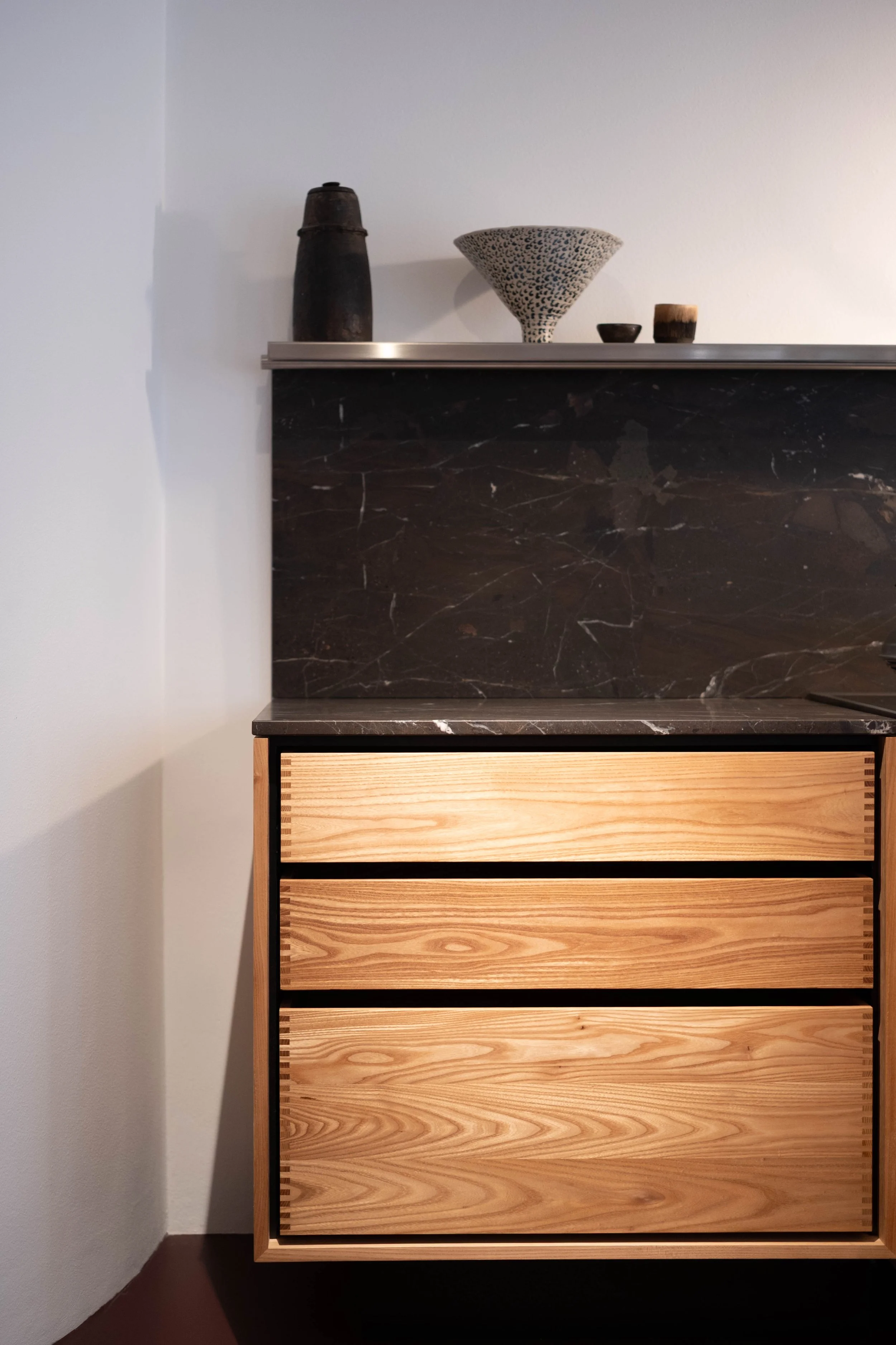 A modern wooden dresser with three drawers, featuring natural wood grain and black framing, placed against a white wall. On top of the dresser are decorative items including a black ceramic vase, a speckled ceramic bowl, and two small wooden cups.