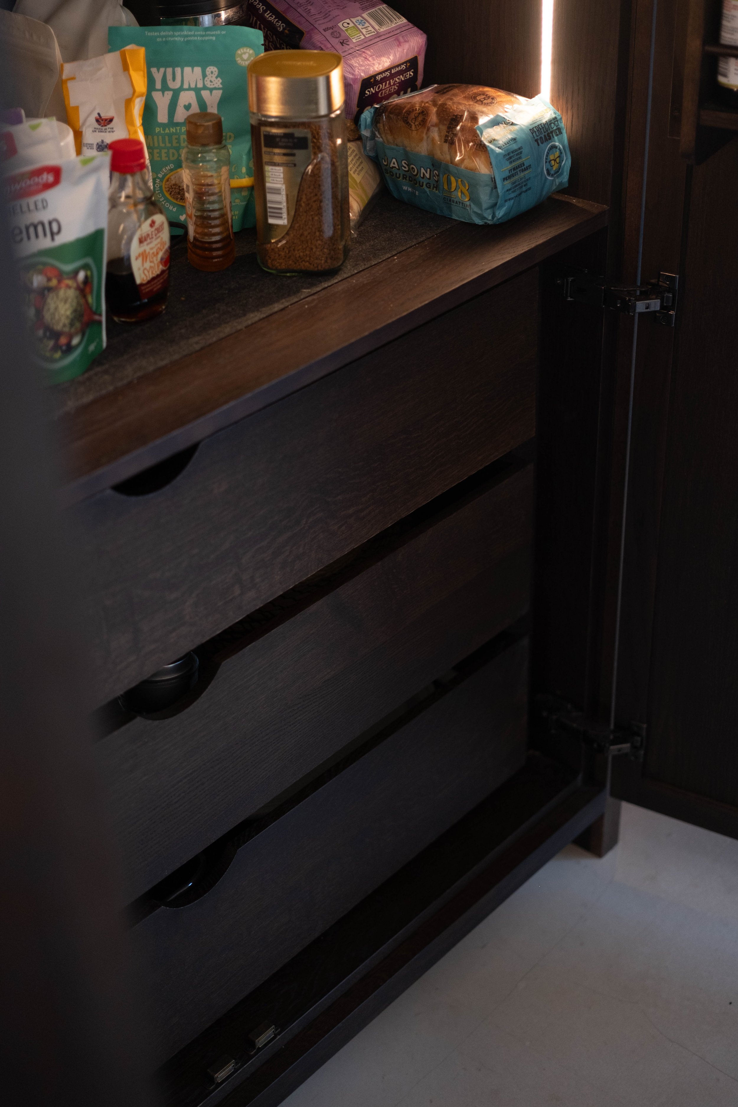 Open dark wood kitchen cabinet with various food and spice items on the shelf, including bread, sauces, and seasonings.
