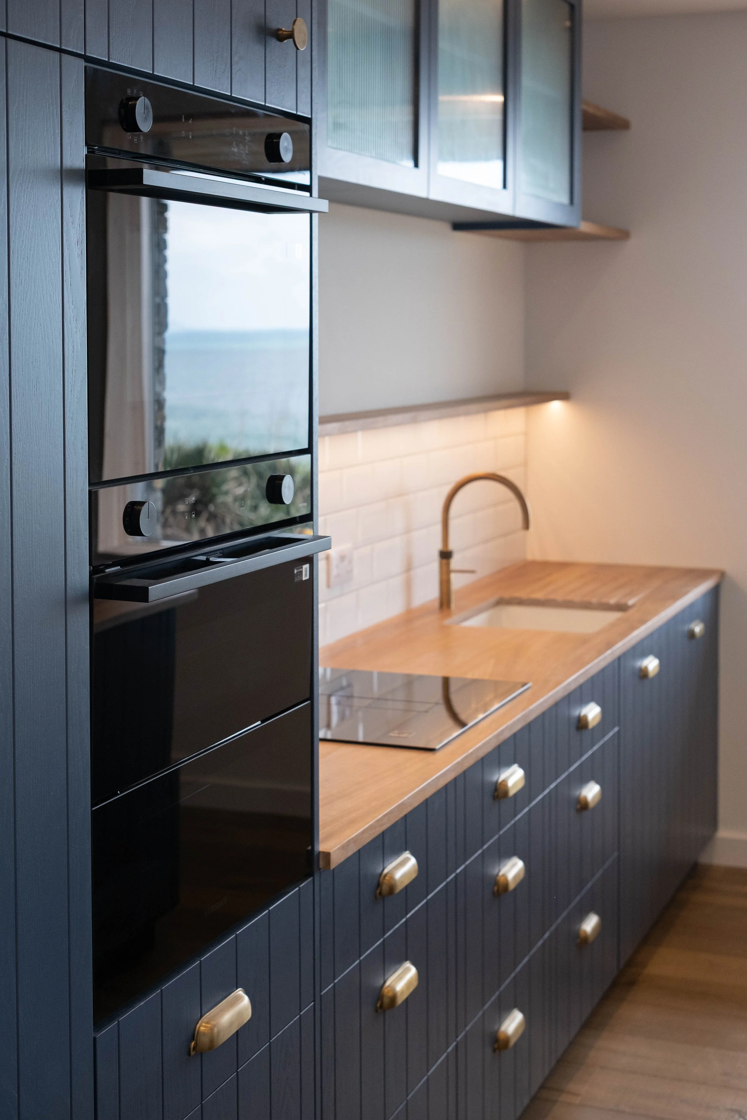 Modern kitchen with dark blue cabinets, wooden countertops, built-in black oven, white tiled backsplash, brass handles, and a gold-finished faucet over a white sink.