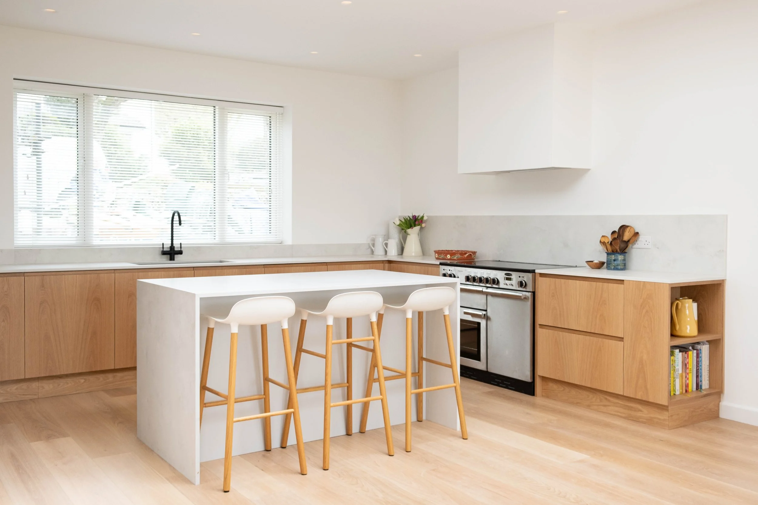 Modern kitchen with large window, white walls, wooden cabinetry, white island with four wooden barstools, black faucet, stove, and plants and utensils on the counter.