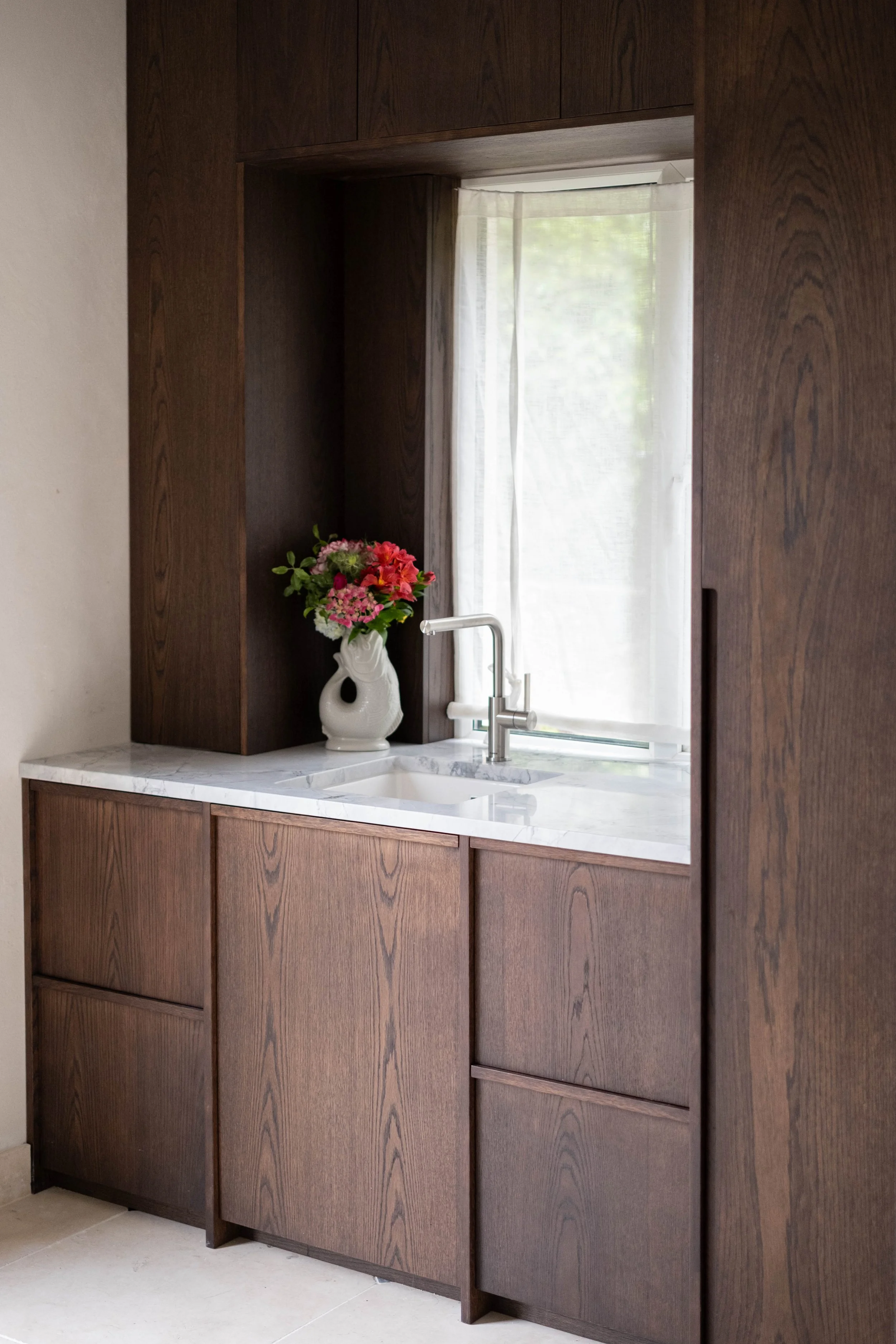Kitchen with dark wood cabinets, a white marble countertop, a white vase with pink flowers, a silver faucet, and a window with sheer curtains.