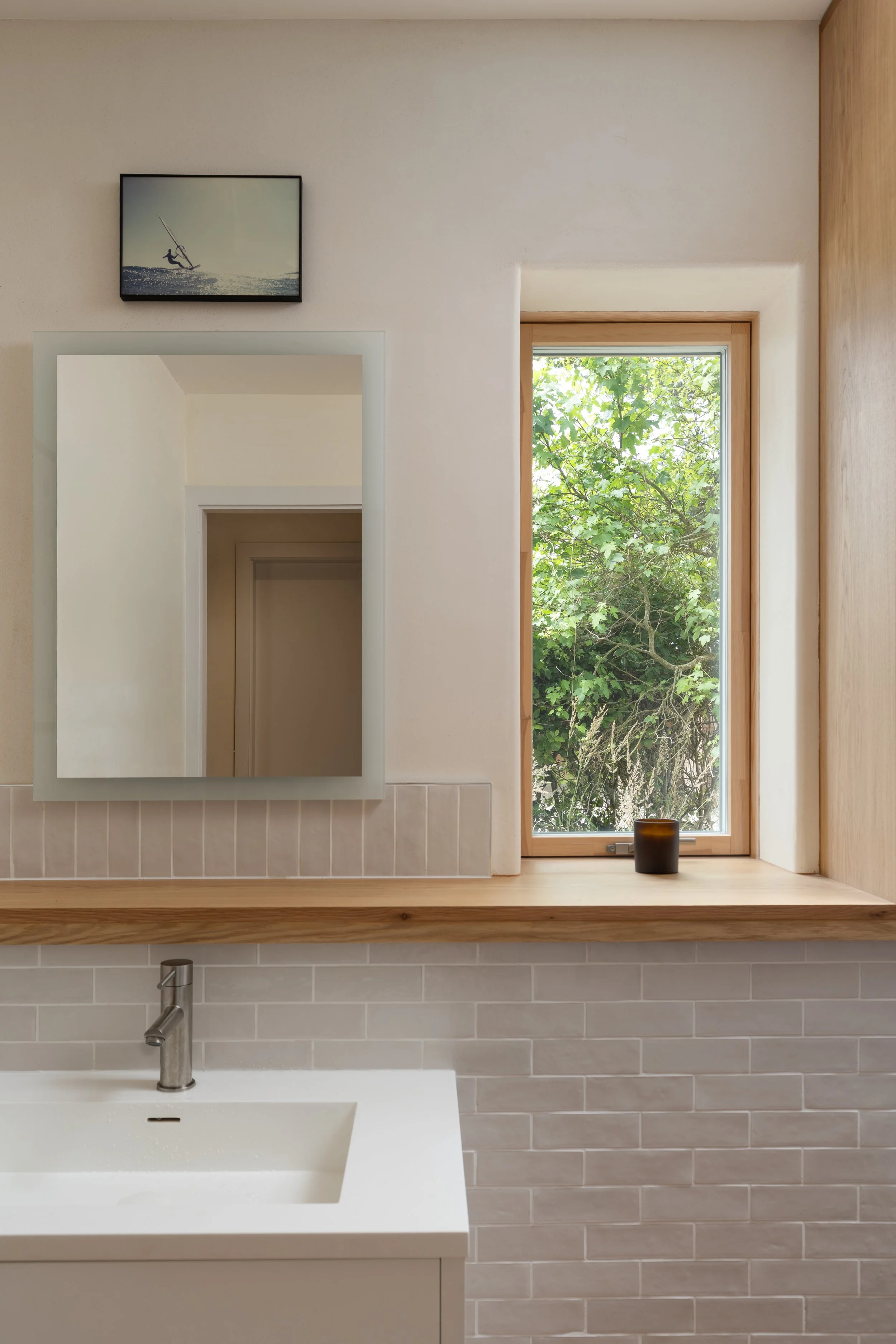 A bathroom with a white sink, a mirror, a wooden ledge, a window with greenery outside, and a small black candle on the ledge.