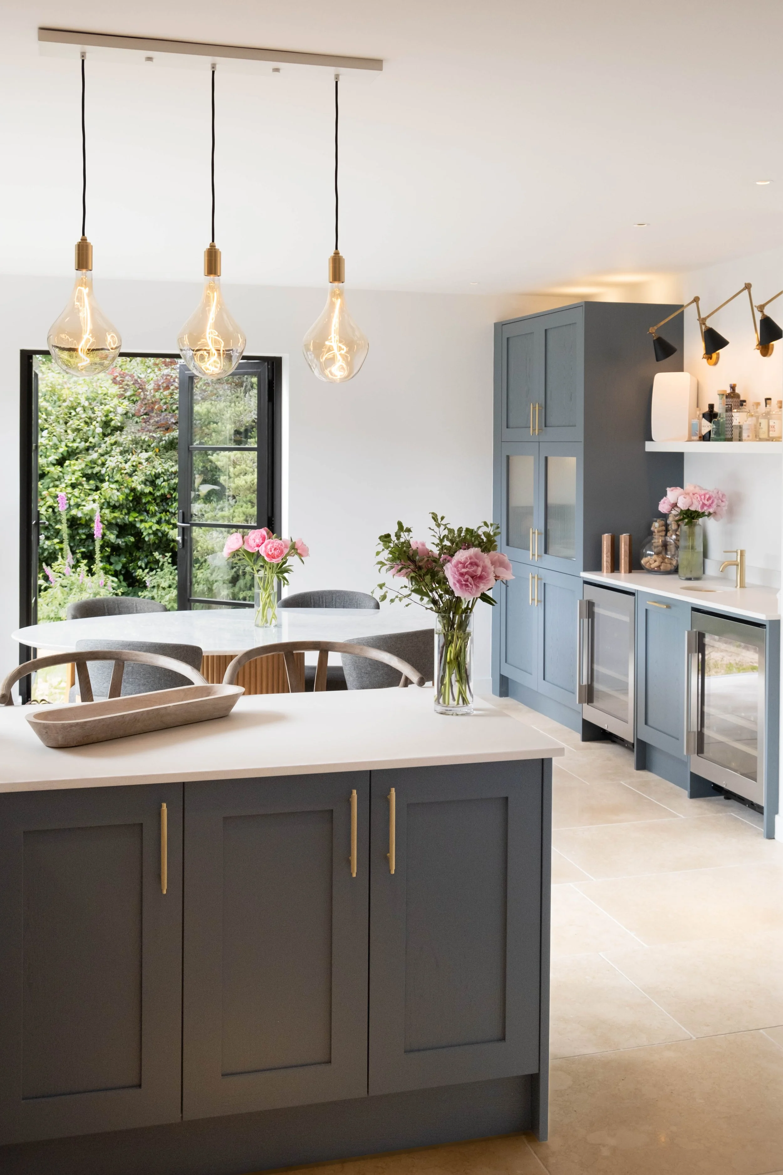 Modern kitchen with blue cabinets, white countertops, a white farmhouse sink, a white table with a vase of pink flowers, and black pendant lights with Edison bulbs hanging from the ceiling. Large window with a view of a garden.
