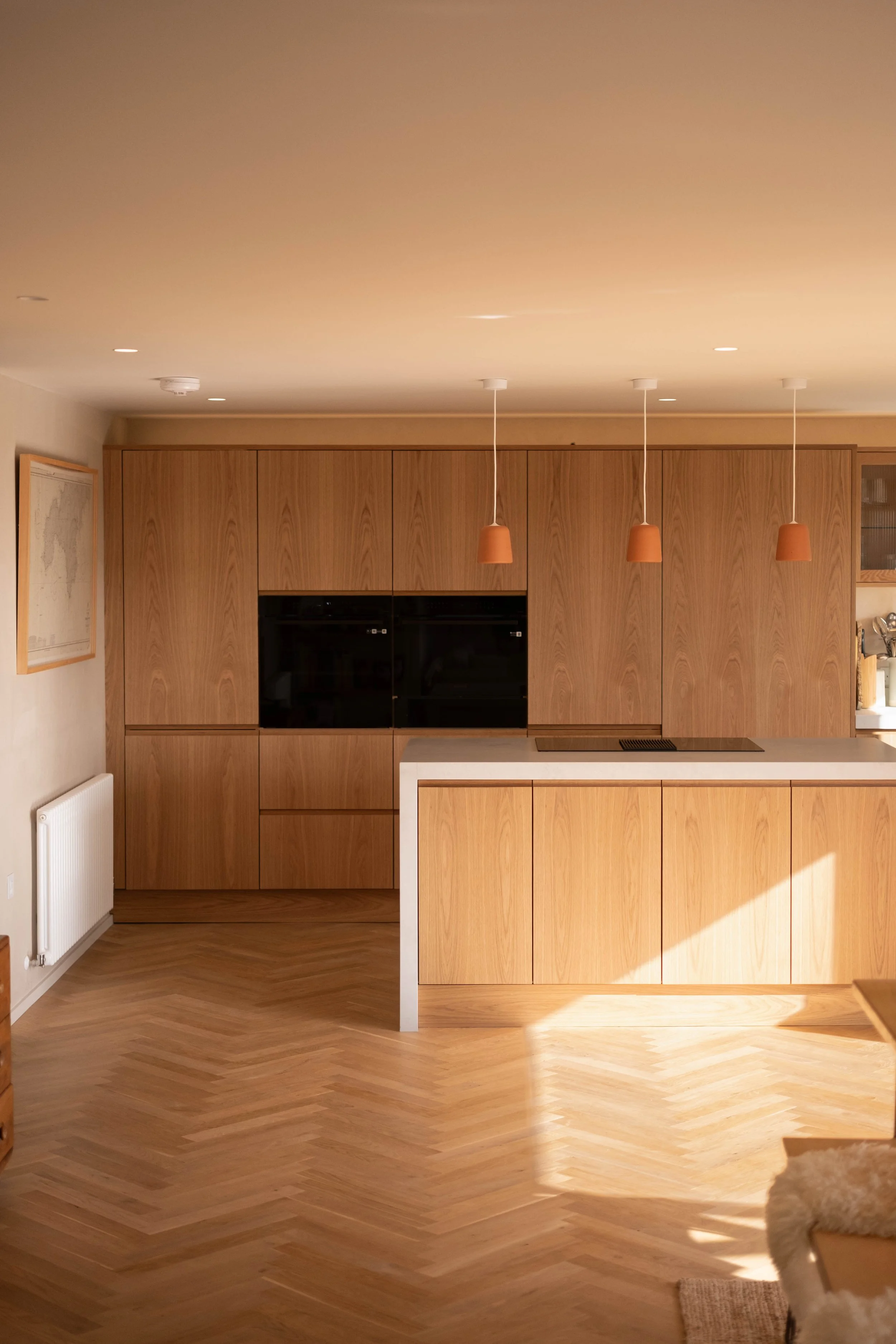 A modern kitchen with wooden cabinets, a white countertop, three hanging pendant lights with terracotta shades, and a wooden chevron-patterned floor. There is a partially visible beige and white fluffy dog in the lower right corner.