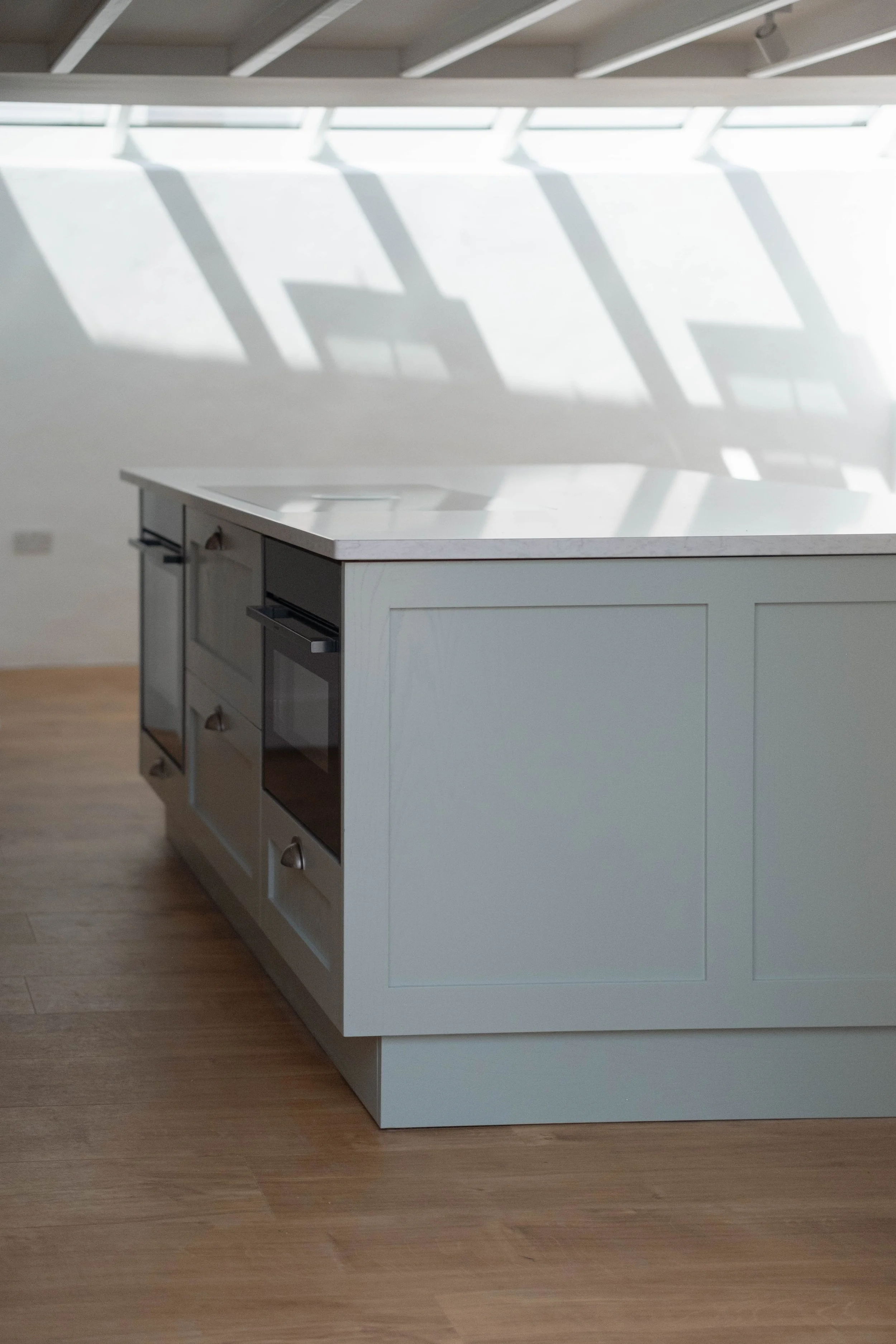 A white kitchen island with a marble top and several drawers and cabinets, situated on a wooden floor in a bright room with large slanted windows and shadows on the wall.