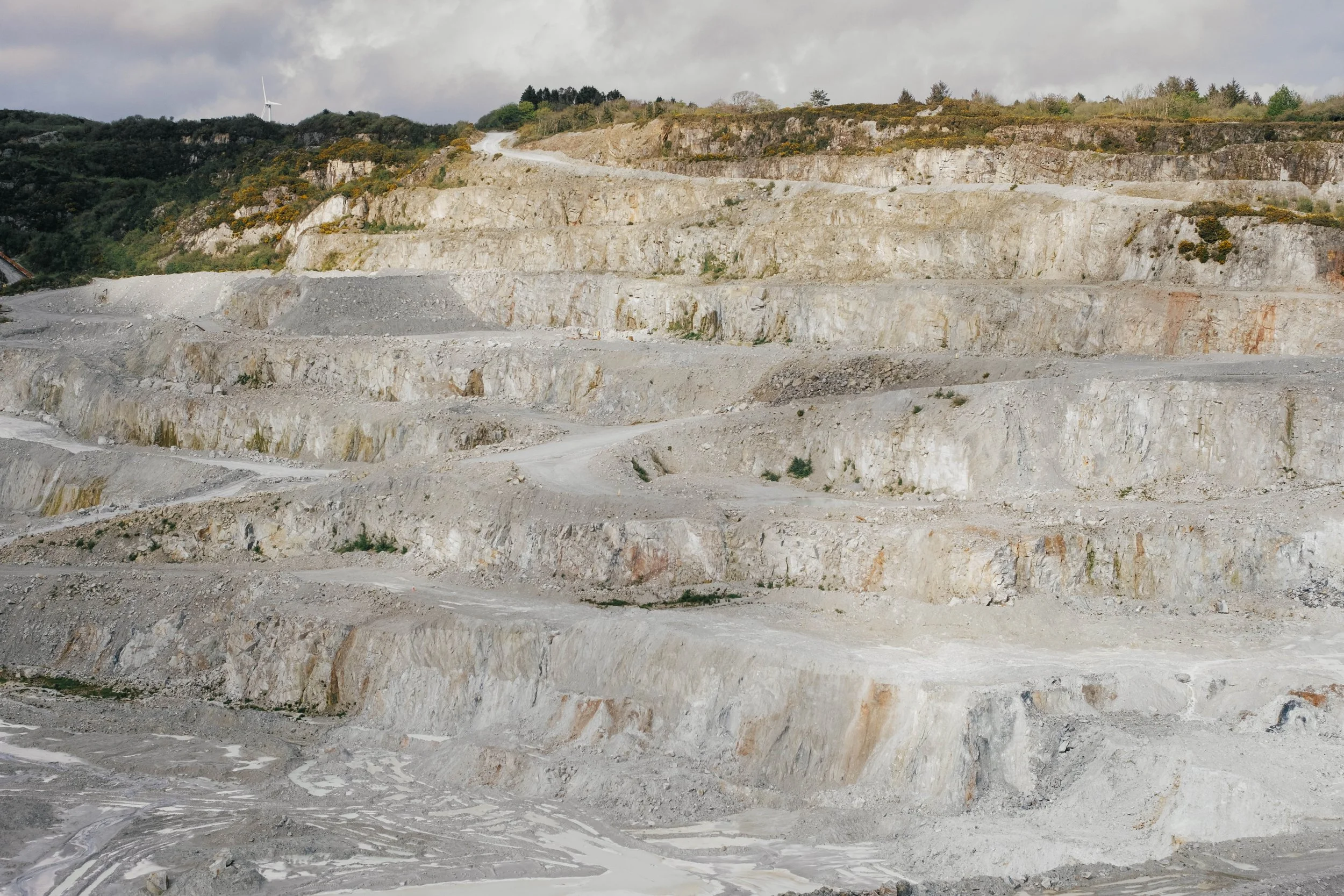 A landscape of a terraced quarry with exposed rocky layers and a winding dirt road, a wind turbine visible on the hilltop in the background under cloudy sky.