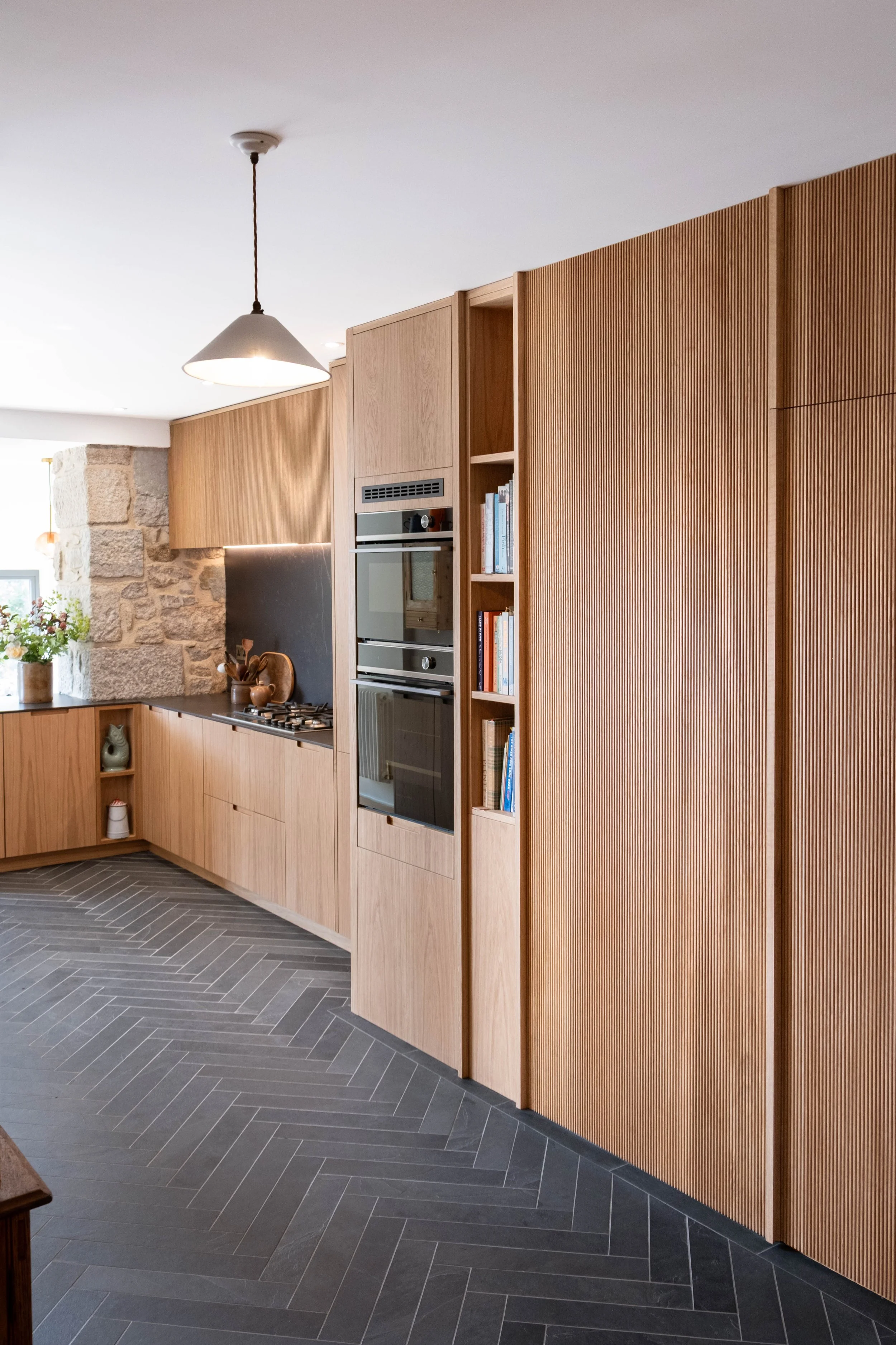 Modern kitchen with wooden cabinets, built-in oven, stone wall, plants, and dark herringbone tile flooring.