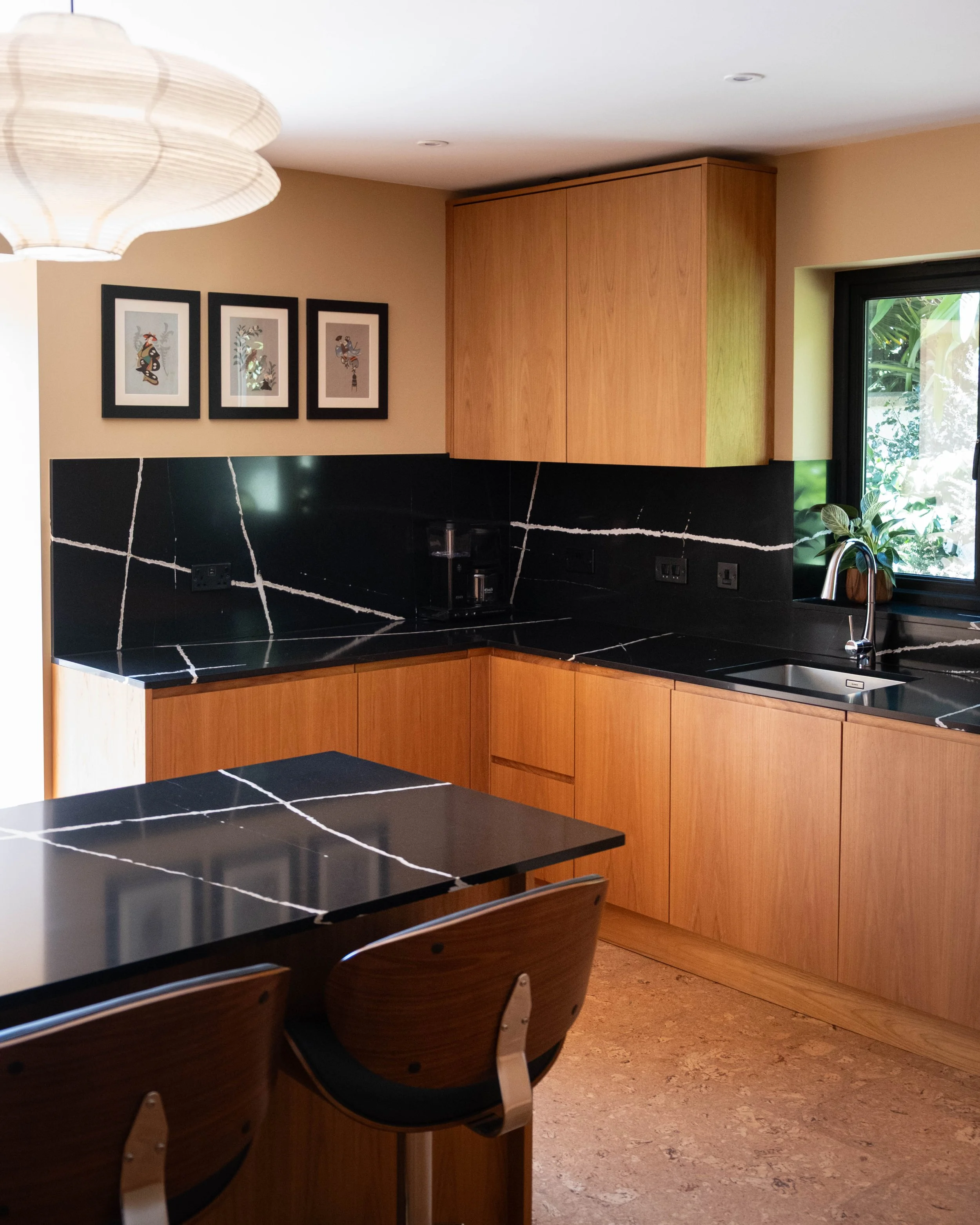 Modern kitchen with black marble countertops with white veining, wooden cabinetry, a window showing outdoor greenery, and three art pieces on the wall.