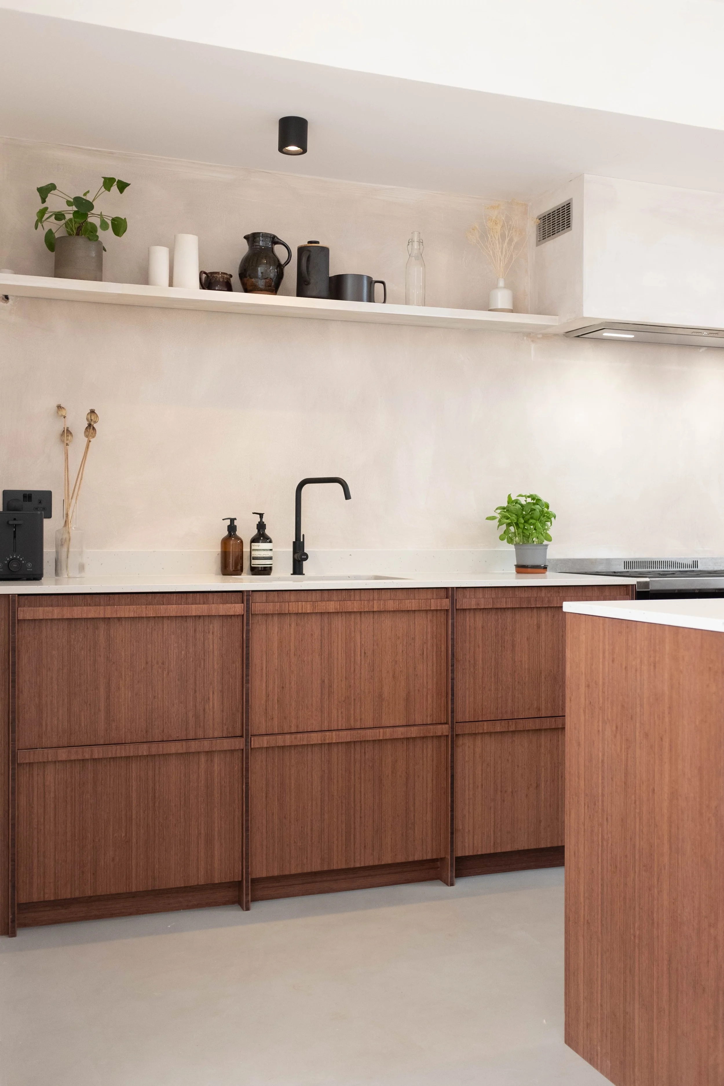 A kitchen with a white countertop, wooden cabinets, and a black faucet. Decor includes a small potted green plant, brown and black bottles, soap dispensers, and vases with dried plants and a green plant.