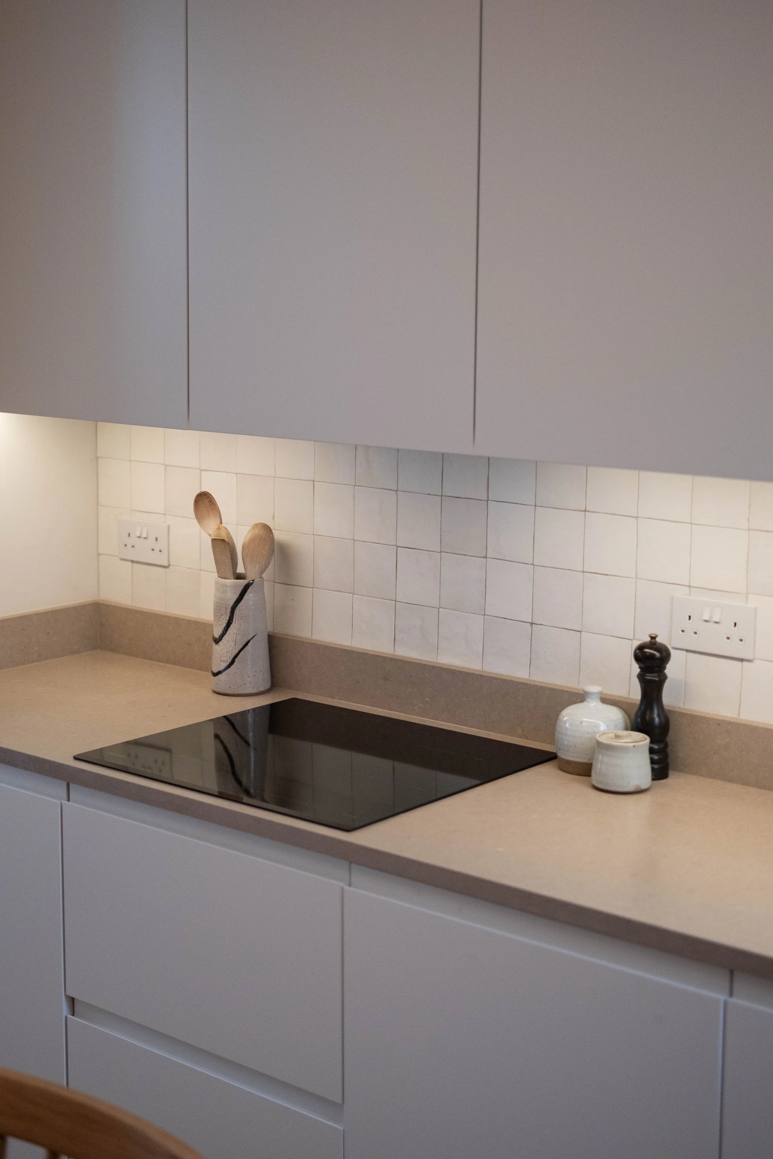 Modern kitchen with white cabinets, beige countertop, black cooktop, decorative vases, and a utensil holder with wooden spoons.