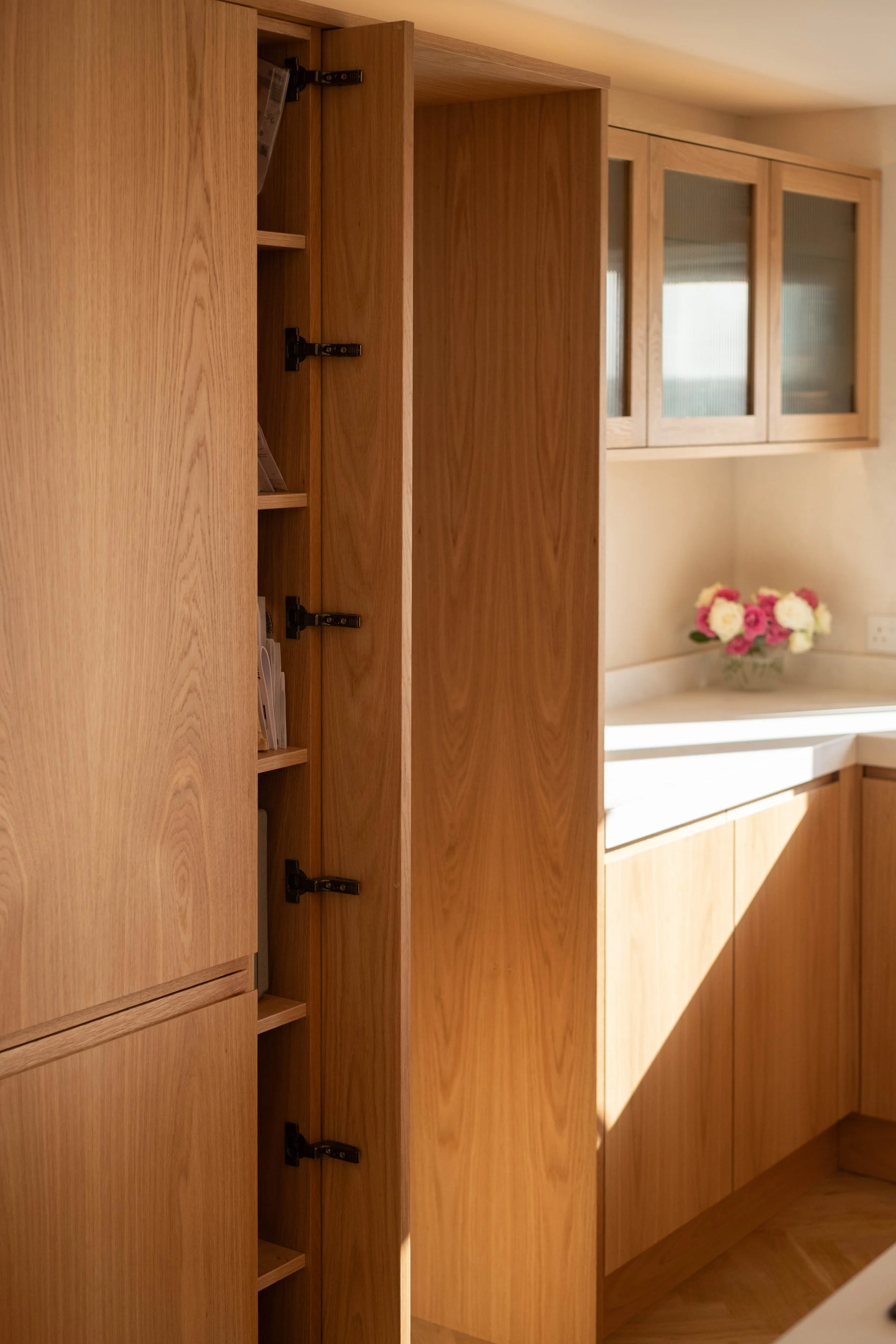 A wooden kitchen cabinet with open shelves and a window with three glass panes above the counter, and a bouquet of pink and white flowers on the countertop.