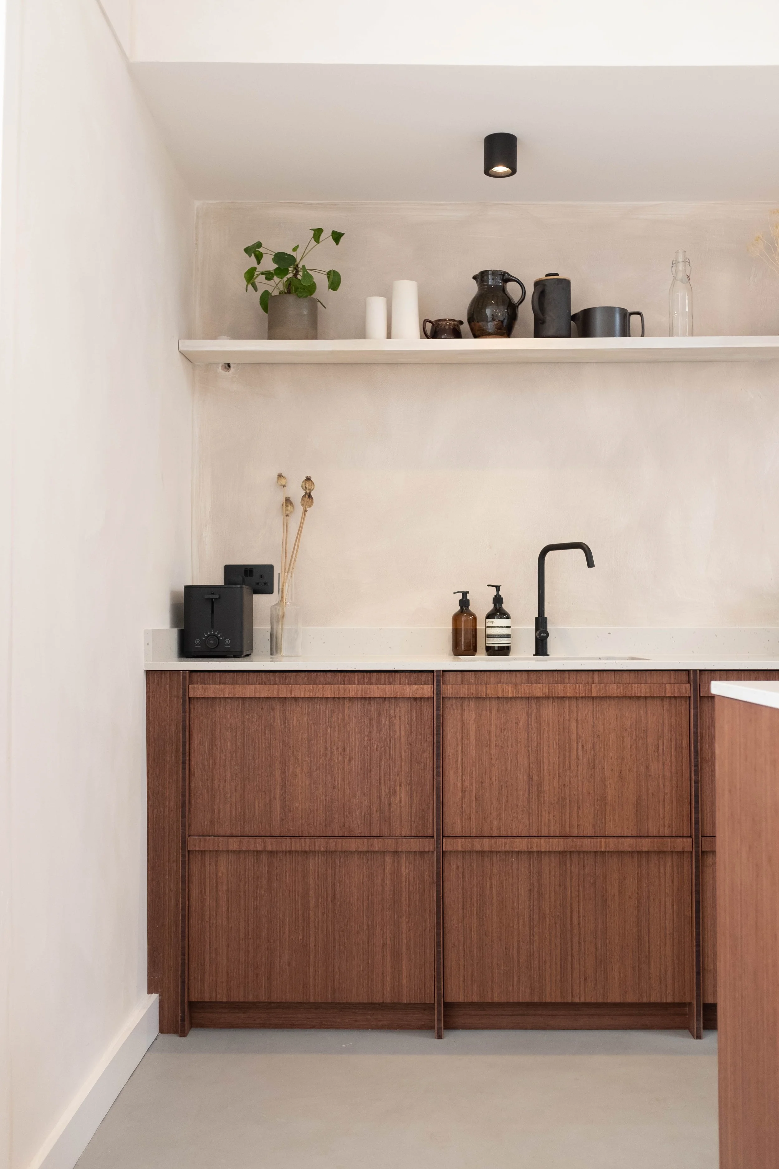Kitchen with wooden cabinets, black faucet, soap dispensers, a white countertop, a shelf with decorative items, a potted plant, vases, pitchers, and a bottle, under a black ceiling light.