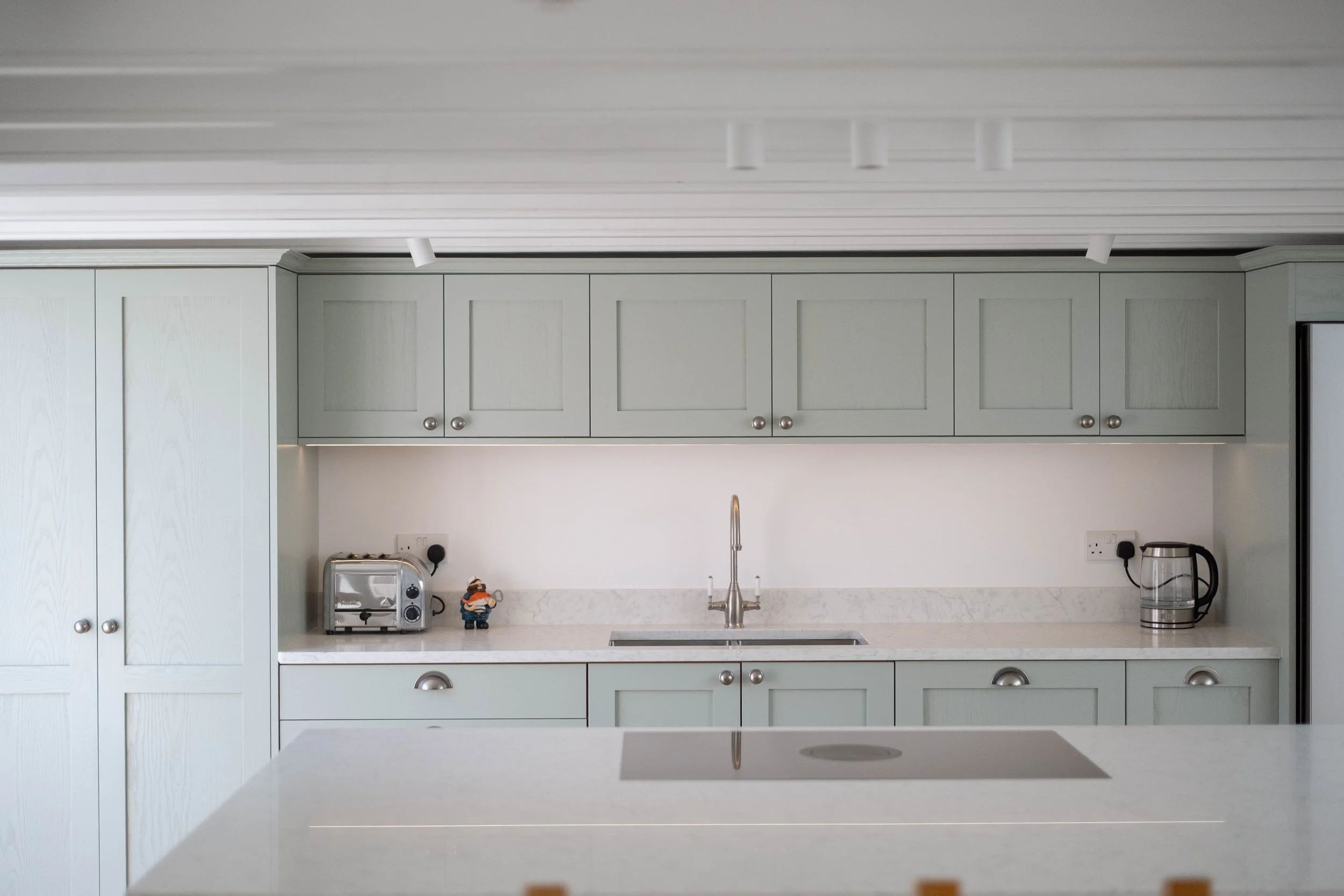A kitchen with light green cabinets, a white marble countertop, a subway tile backsplash, a silver faucet, small appliances including a toaster and kettle, and decorative figurines.