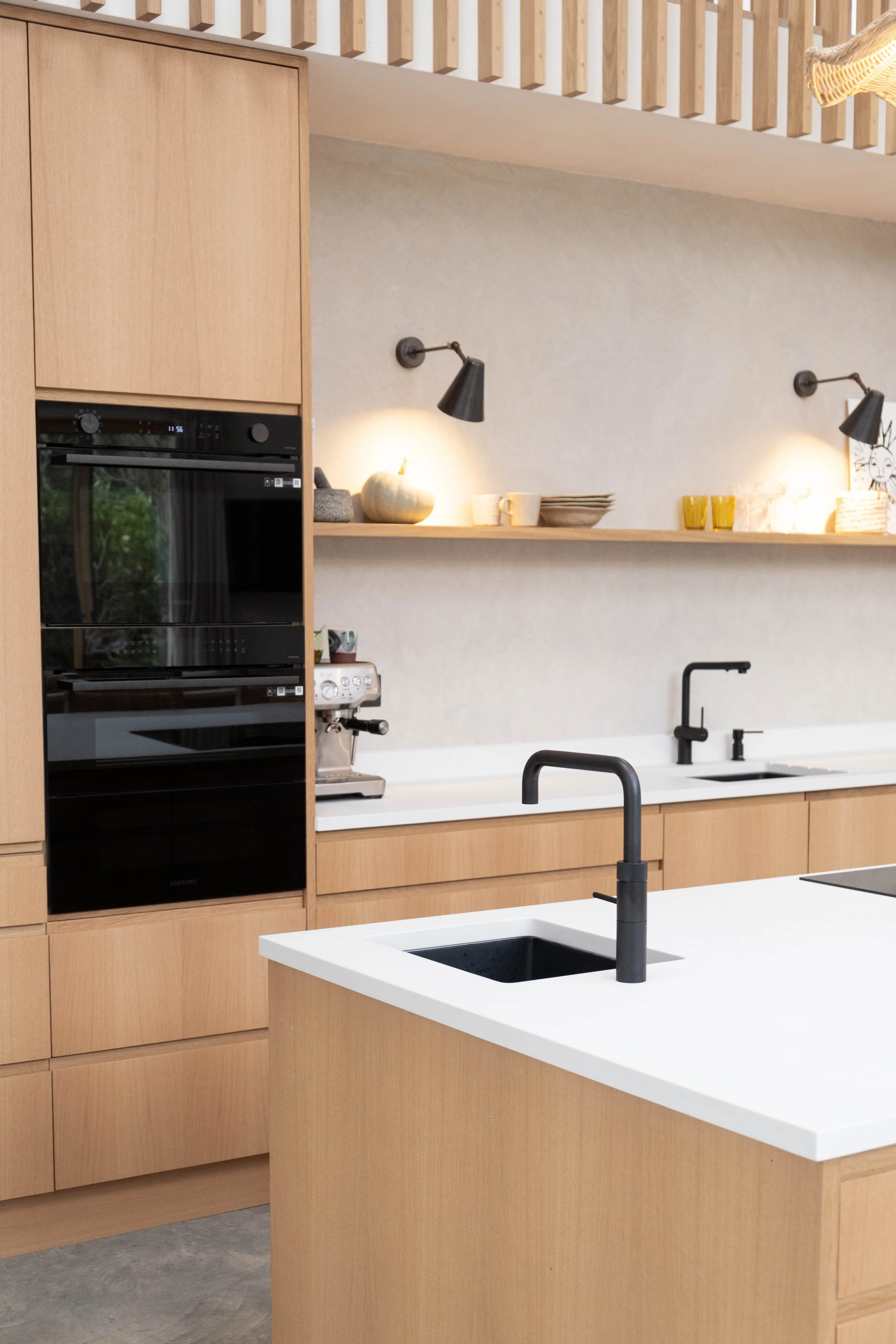 Modern minimalist kitchen with beige wooden cabinets, a white kitchen island with black sink and matte black faucet, black wall-mounted lamps, and open shelves with decorative items and dishware.