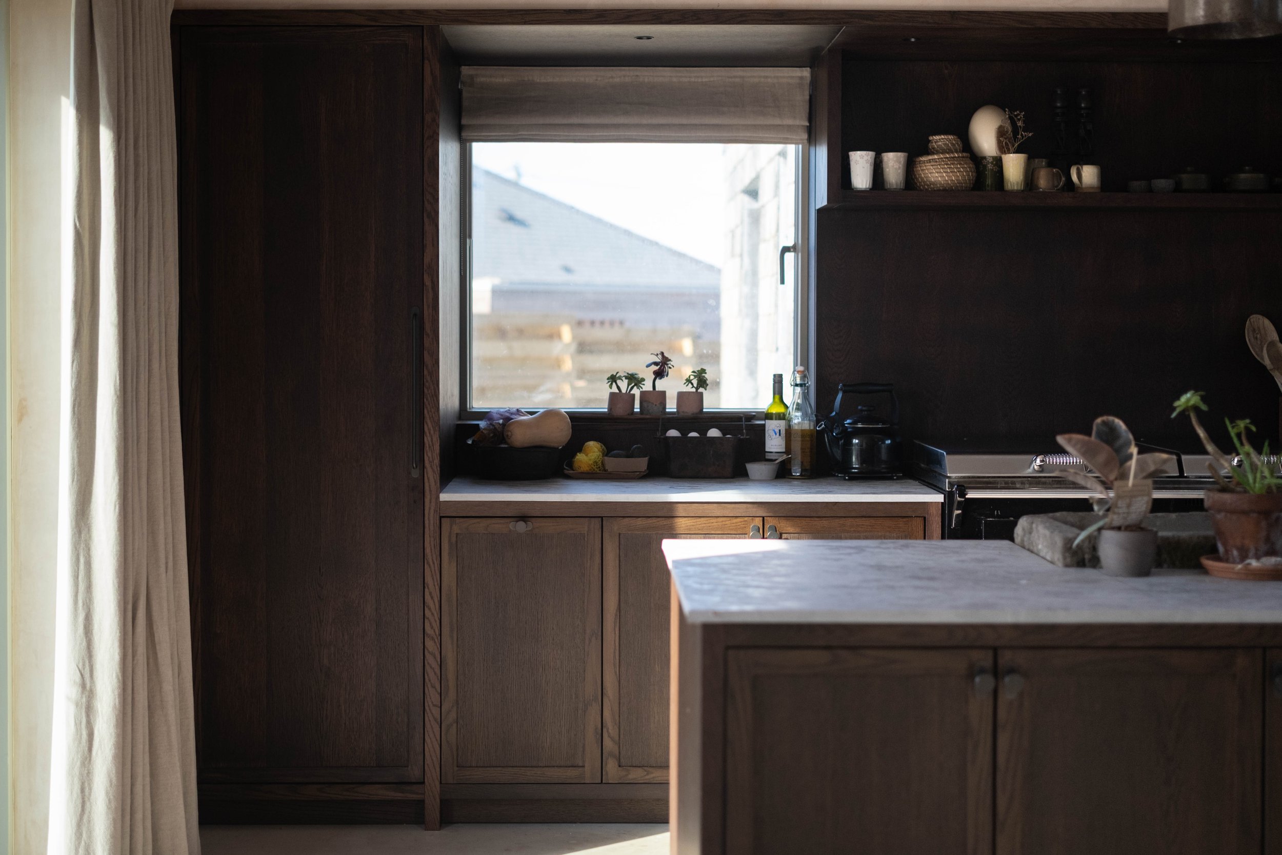A kitchen scene showing a window above the sink with a view of a house roof and blue sky outside. The kitchen has dark wood cabinets and shelves, with various dishes, cups, and decorative items on the shelves. The windowsill has small potted plants, 