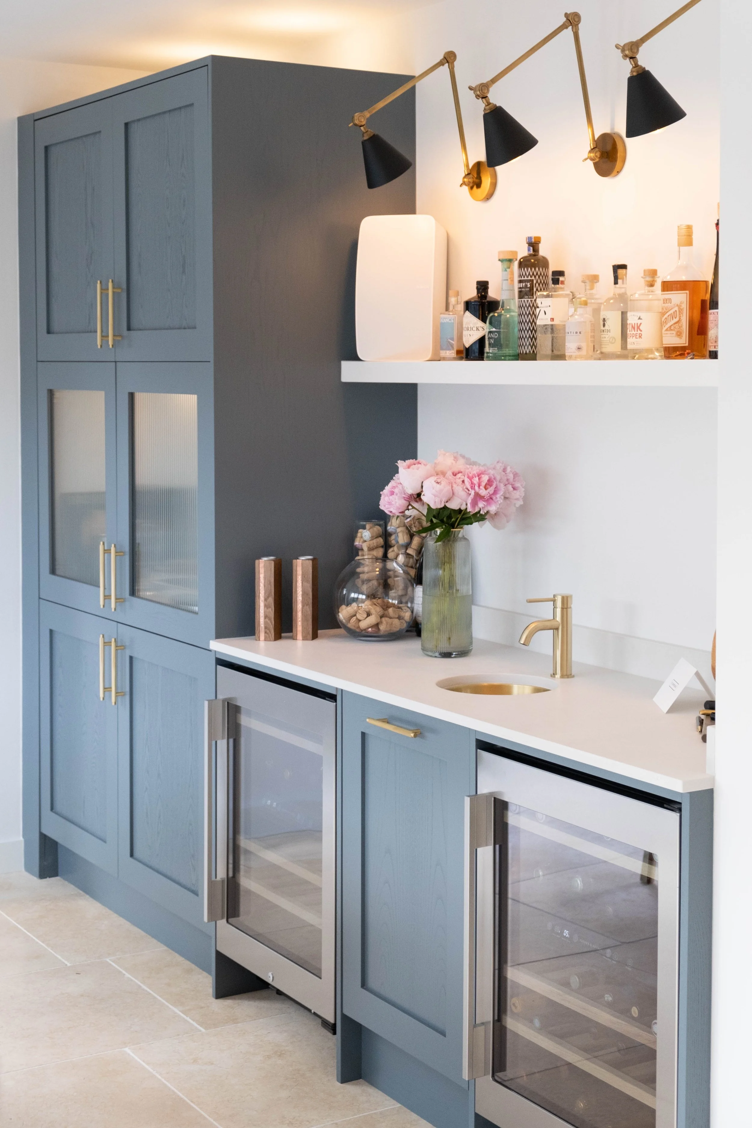 A modern kitchen with blue cabinets, a white countertop, gold handles, and a built-in wine fridge. There are pink flowers in a glass vase, bottles of alcohol on a shelf, and black wall-mounted lamps with gold accents above the shelf.