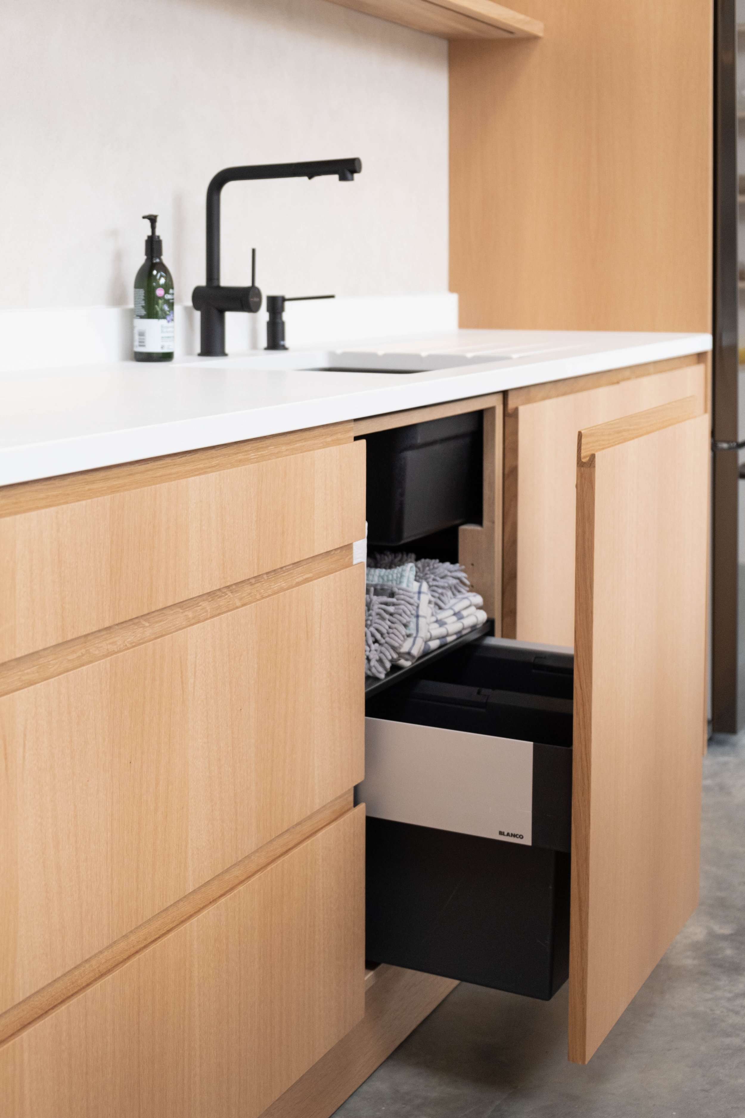Open kitchen cabinet with wooden drawers, a white countertop, a black dishwasher, and a black trash bin underneath. Small bottle of liquid soap or sanitizer on the counter, black kitchen sink and faucet.