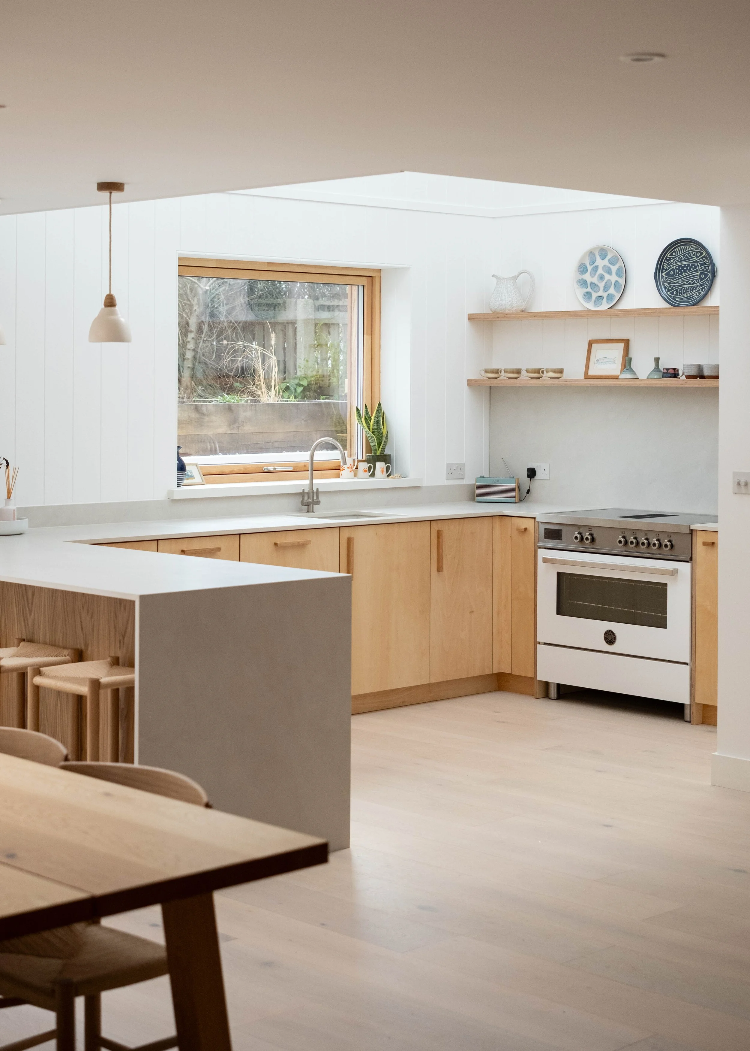 A modern, minimalist kitchen with white walls, wooden cabinets, and a window overlooking a backyard. The kitchen features open wooden shelving with plates and vases, a white range, and a sink under the window. A pendant light hangs from the ceiling, 