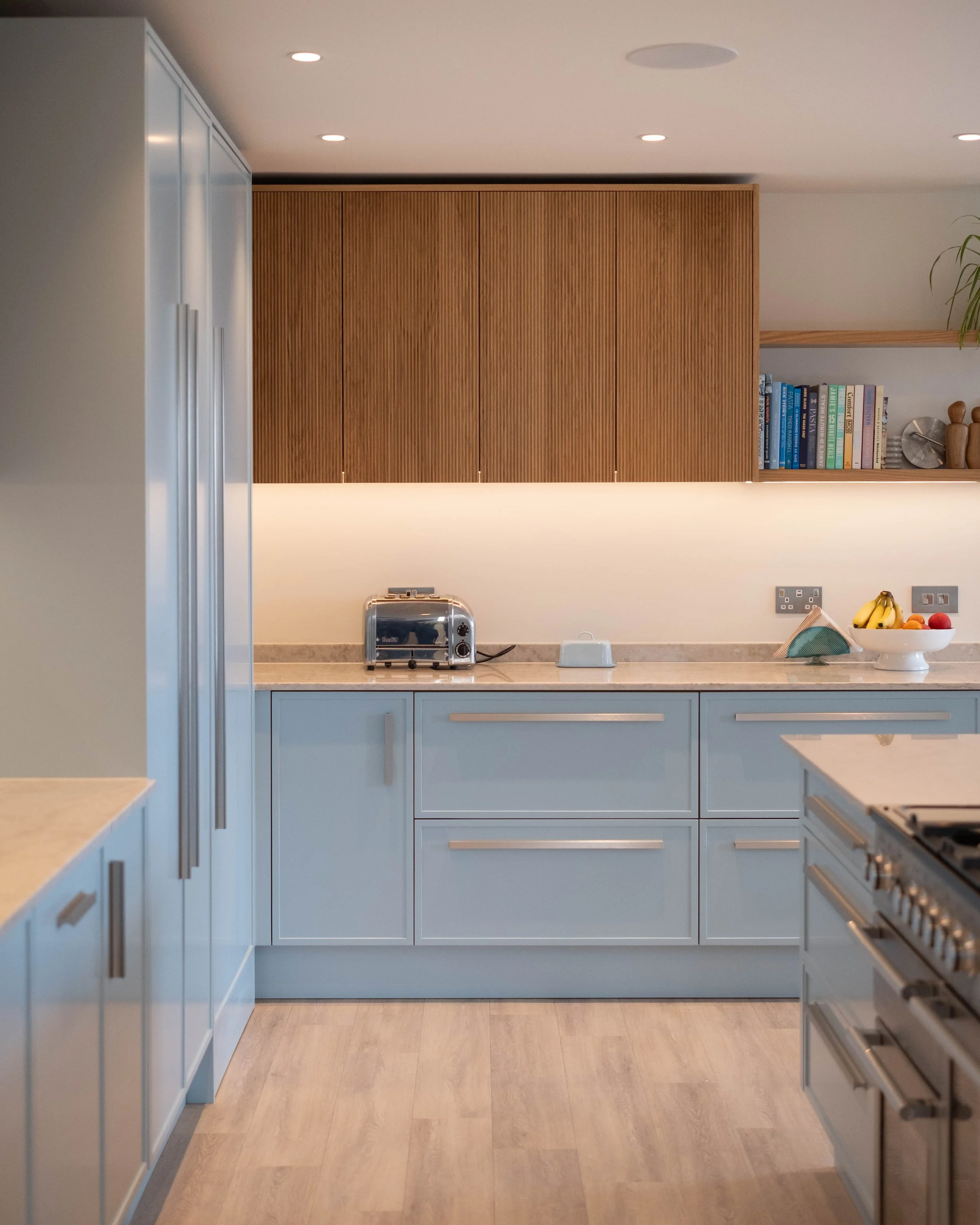 A modern kitchen with blue cabinets and a wooden upper cabinet. Countertop appliances include a toaster oven and a white kettle. Open shelves hold books, plants, and decorative items. Fruit bowl on the counter contains bananas and other fruits. The f