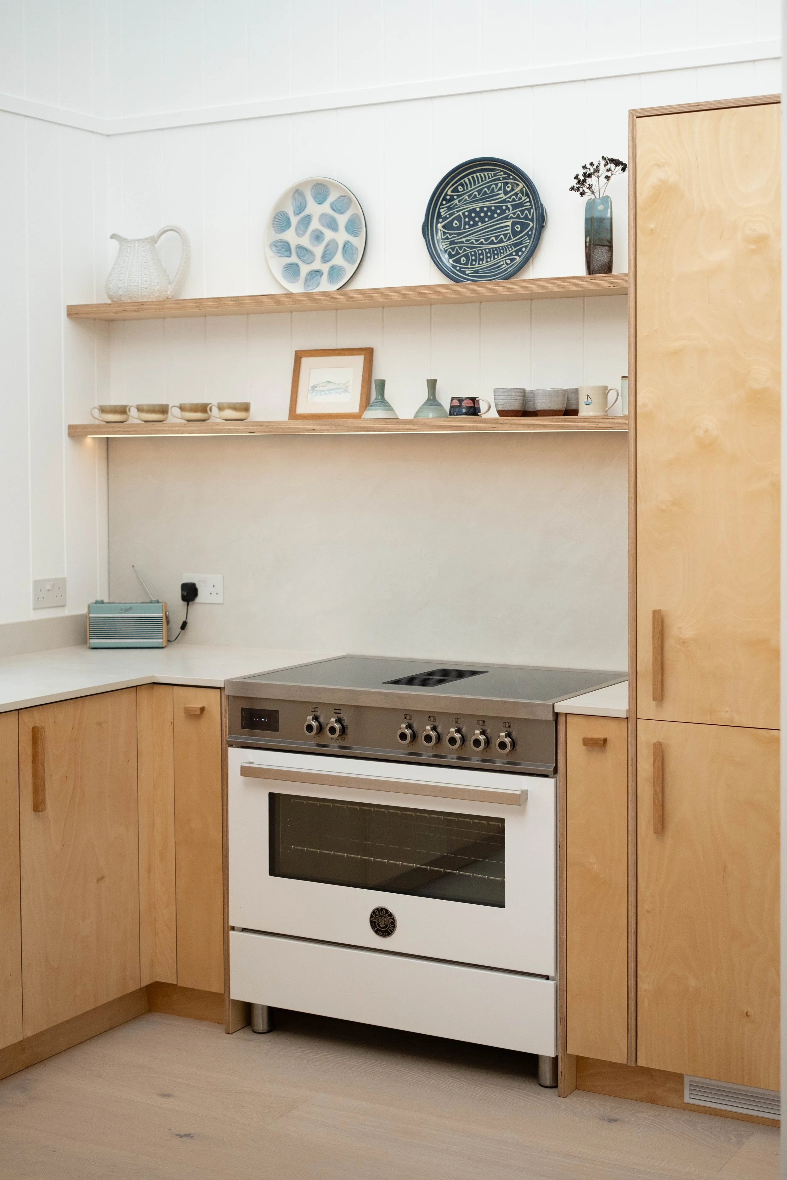 A kitchen with wooden cabinets, a white oven, and black stovetop. Two wooden shelves display various kitchenware, including plates, bowls, cups, and a framed picture. A small blue radio is on the counter, and a vent is near the floor.