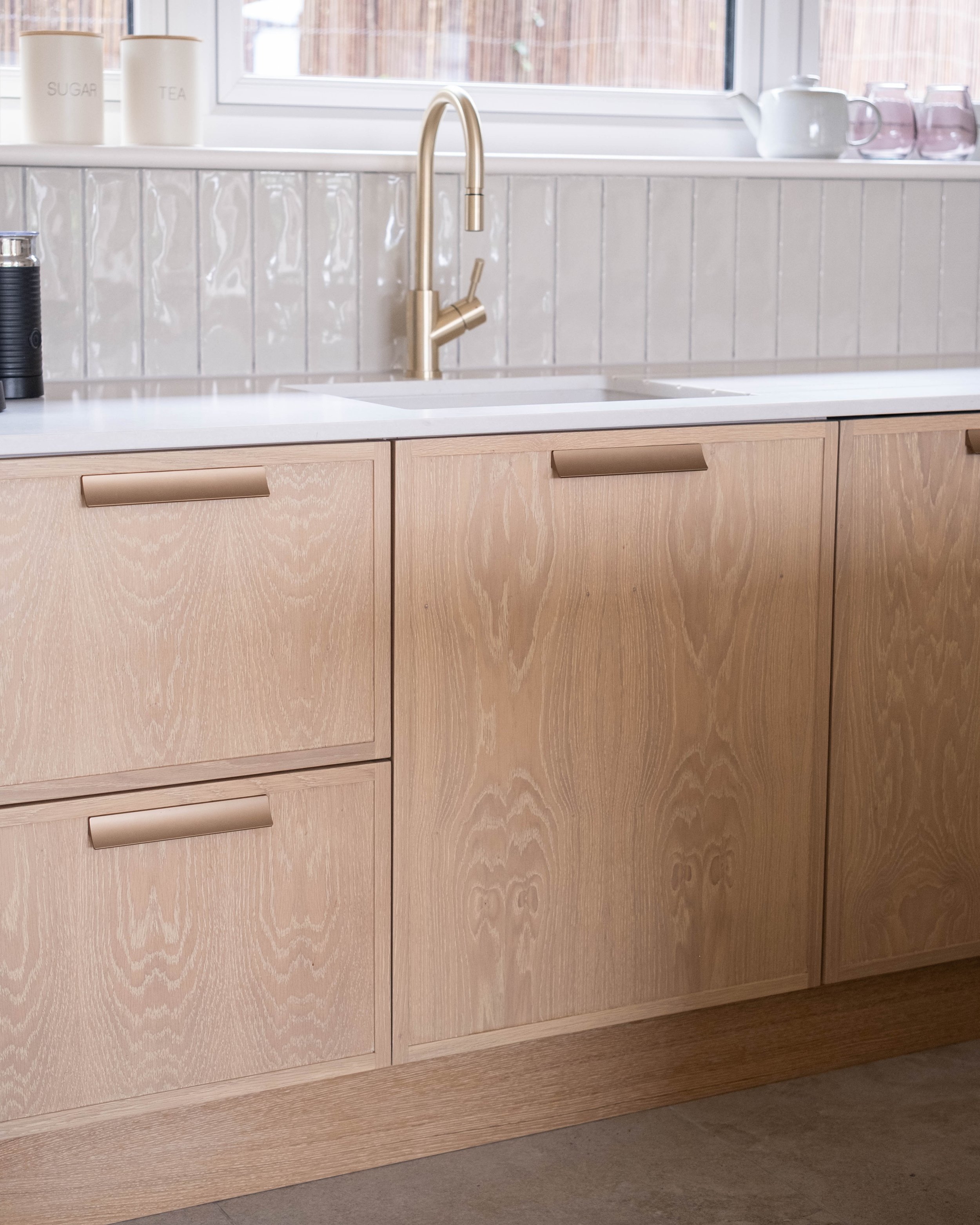 A kitchen sink with a gold faucet, wooden cabinets, and a window in the background. On the windowsill, there are three white containers labeled 'SUGAR,' 'TEA,' and an unlabeled container, along with a teapot and glasses.