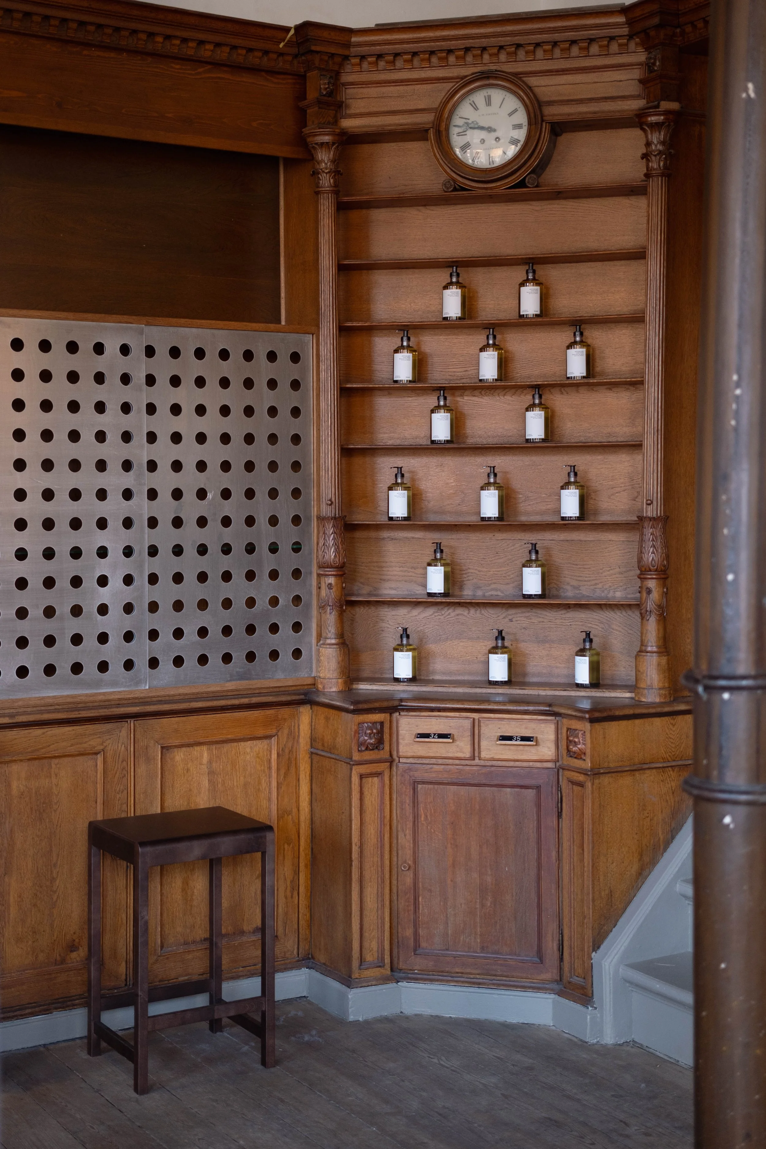 A vintage wooden pharmacy or apothecary cabinet with multiple shelves filled with empty bottles, a round clock showing 10:11, and a small stool nearby.