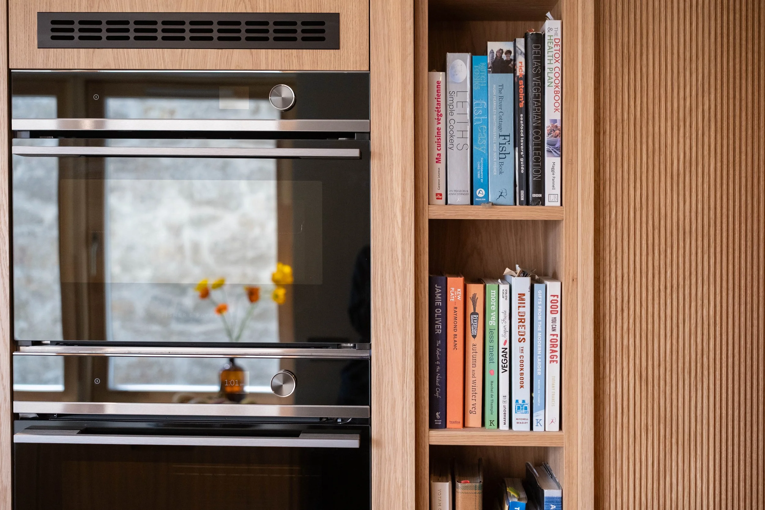 A kitchen with a built-in oven, glass-door microwave, and a wooden bookshelf filled with cookbooks and magazines.