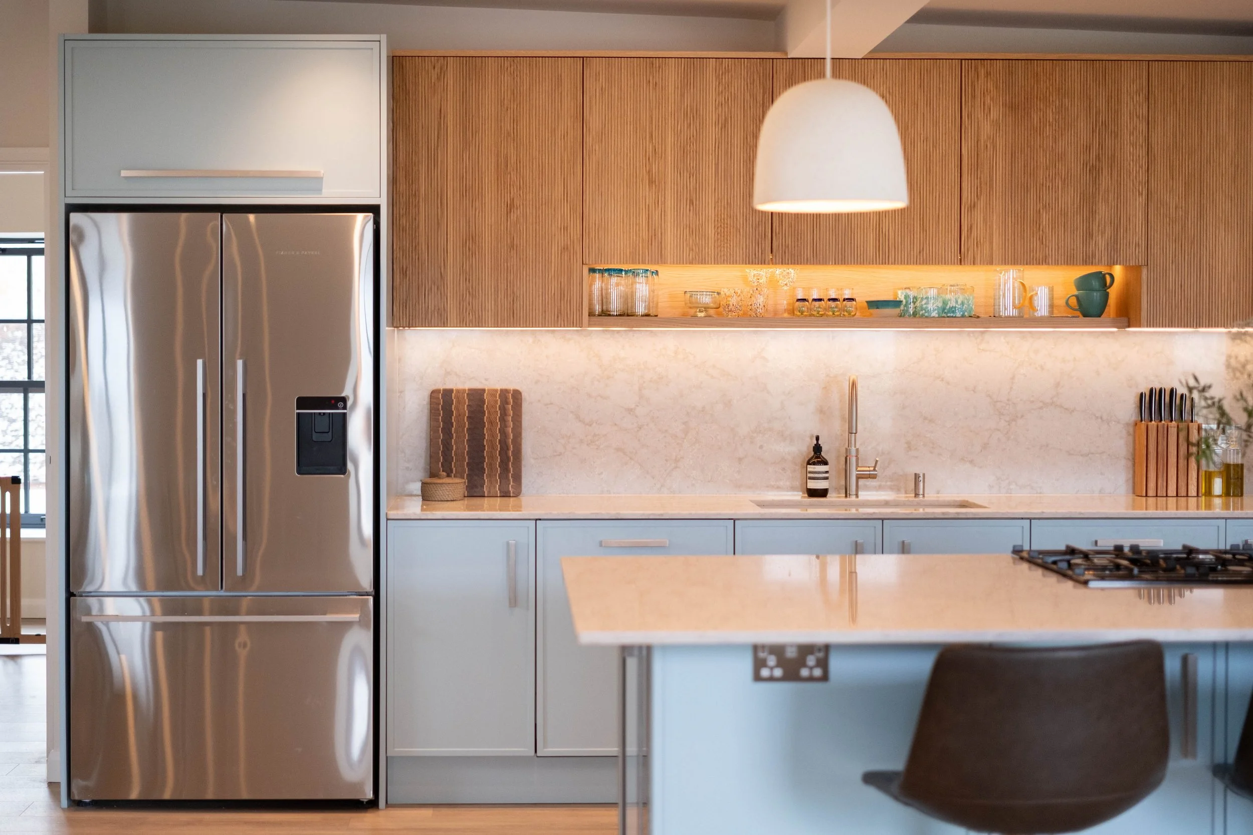 Modern kitchen with stainless steel refrigerator, wooden cabinets, light-colored marble countertop, hanging pendant light, and various glassware and kitchen items.