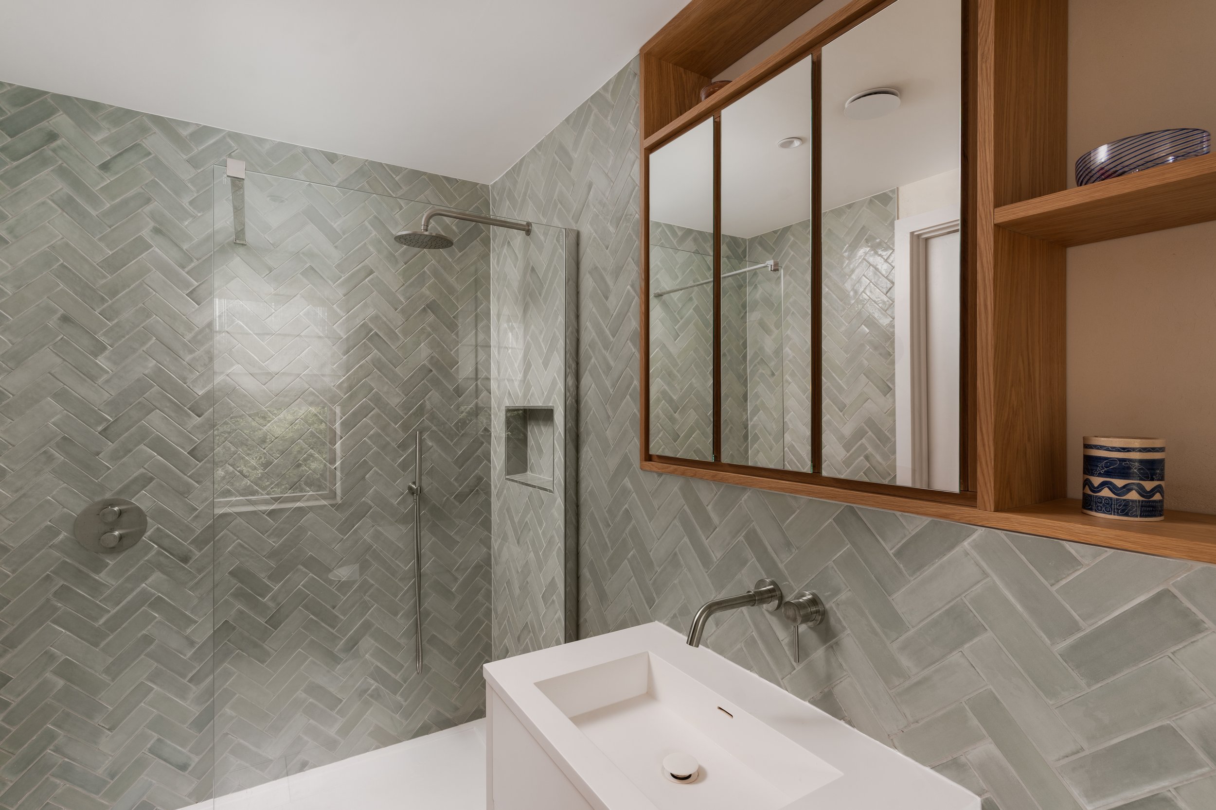 A bathroom with a walk-in shower featuring light green herringbone tile, a rain showerhead, and a small niche for bath products, a wooden-framed mirror, a shelving unit with decorative bowls, and a white vanity with a vessel sink.