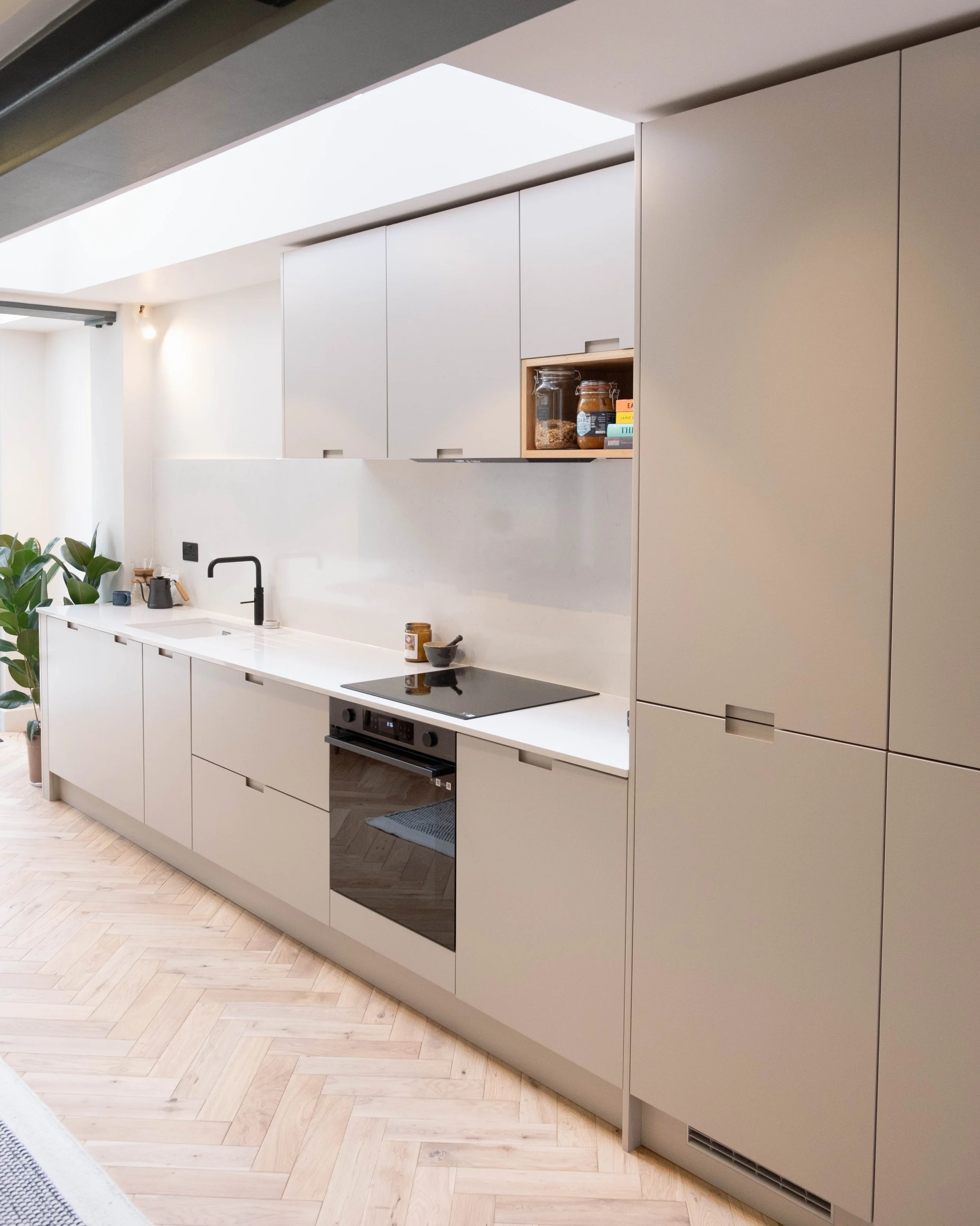 Modern kitchen with beige cabinets, black induction cooktop, built-in oven, wooden flooring, and a corner with potted plants. Skylight visible at the top.
