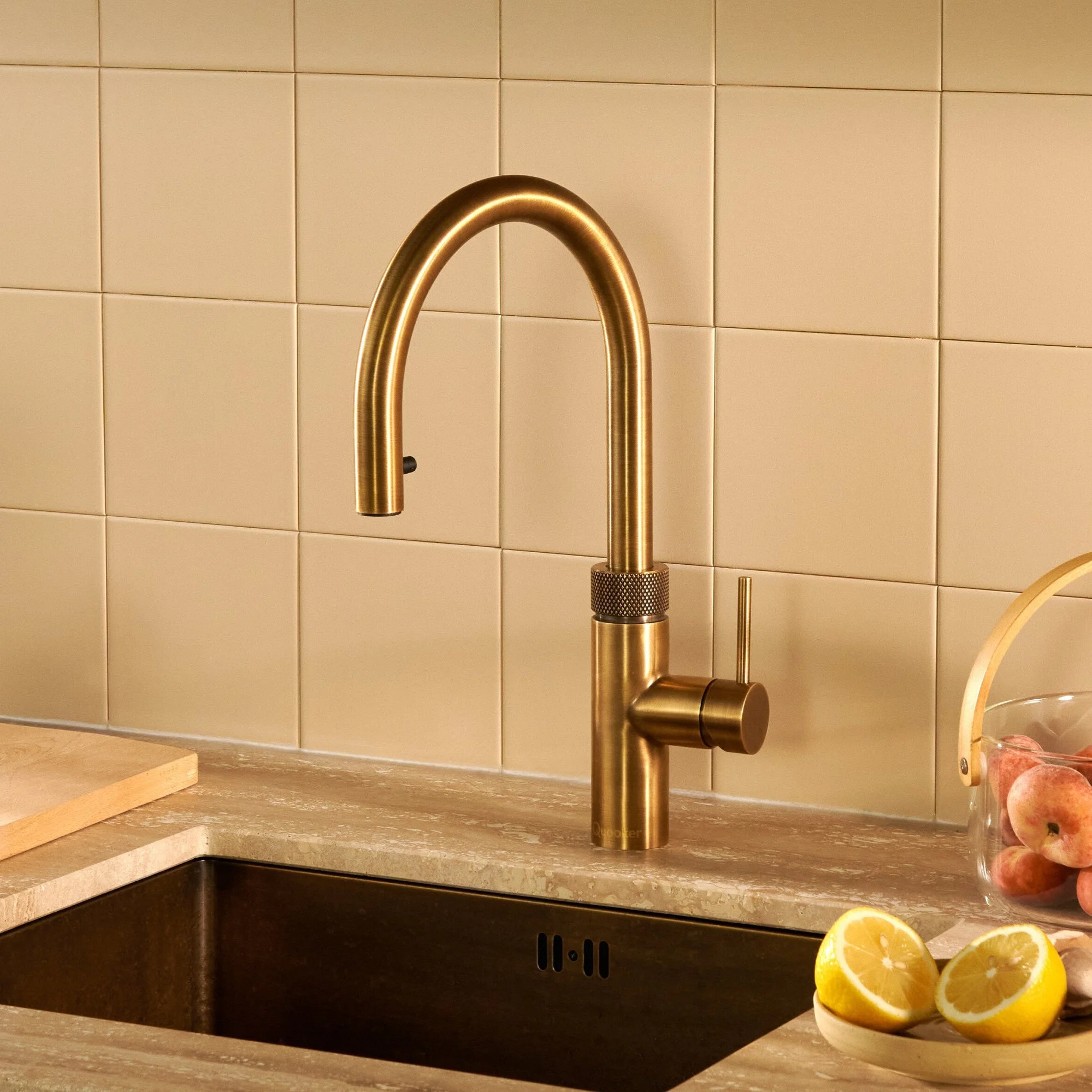 A kitchen faucet on a beige tiled wall above a black sink with a bowl of lemon slices in the foreground.
