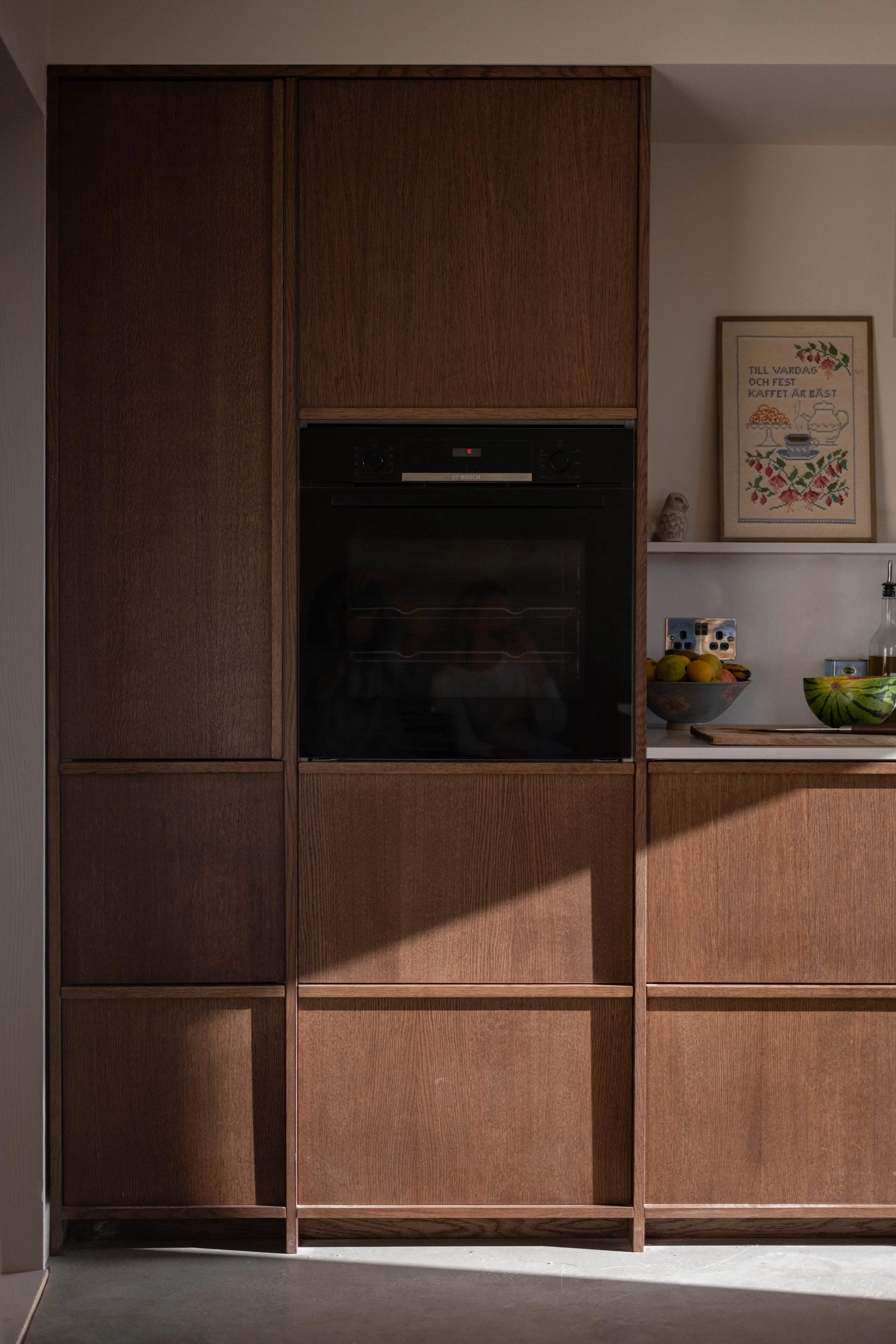 A wooden built-in cabinet with closed compartments and an integrated oven. To the right, a shelf with a framed embroidery piece, bowls with fruits, a bottle, and a textured owl figurine are visible.