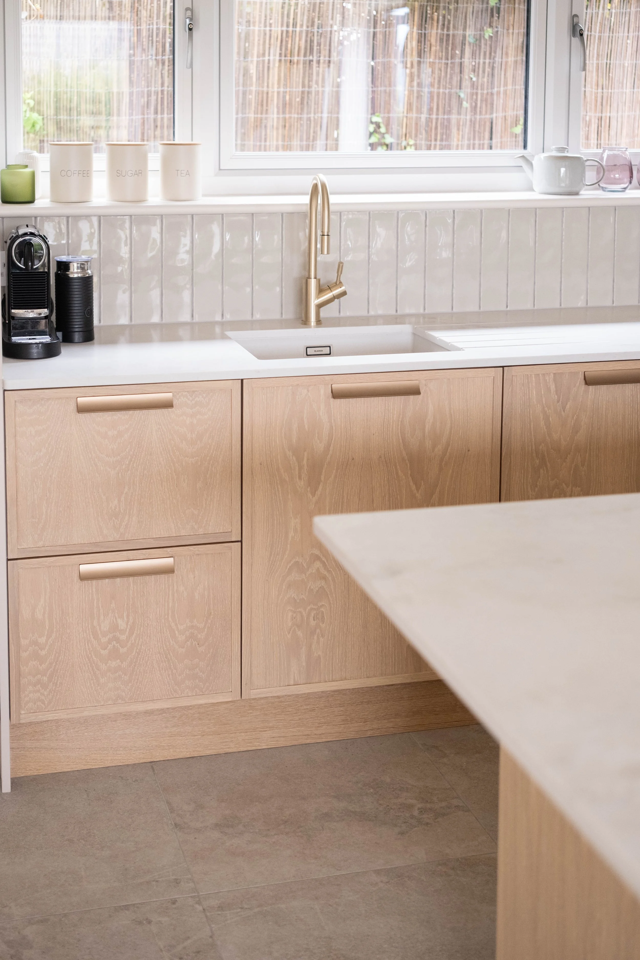 A modern kitchen with wooden cabinets, white countertops, and a stainless steel sink with a gold-colored faucet. On the counter, there is a black coffee machine and a black container. The window above the counter shows an outdoor area with a wooden f