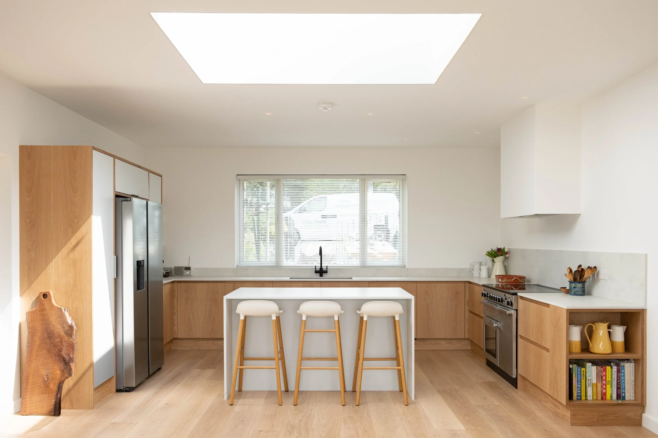 Modern kitchen with wooden cabinets, stainless steel appliances, a white island with three chairs, and a large window with blinds.