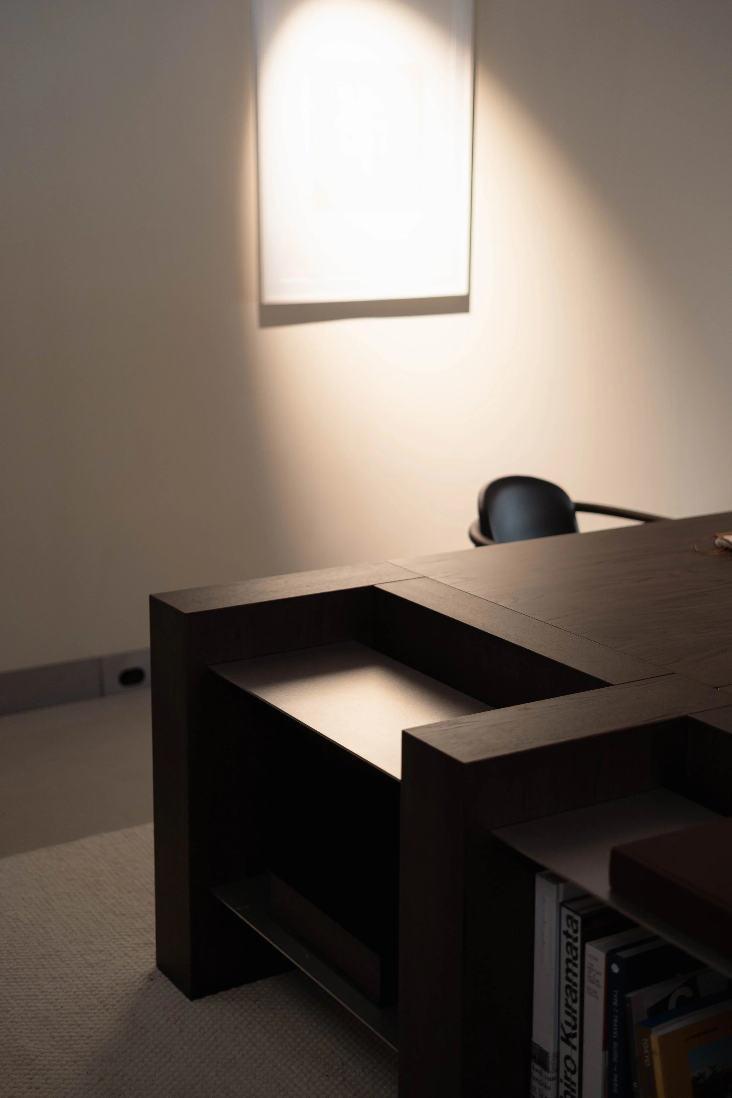 An empty office space with a desk, a black office chair, and a bookshelf filled with books, with a window casting bright light onto the wall.