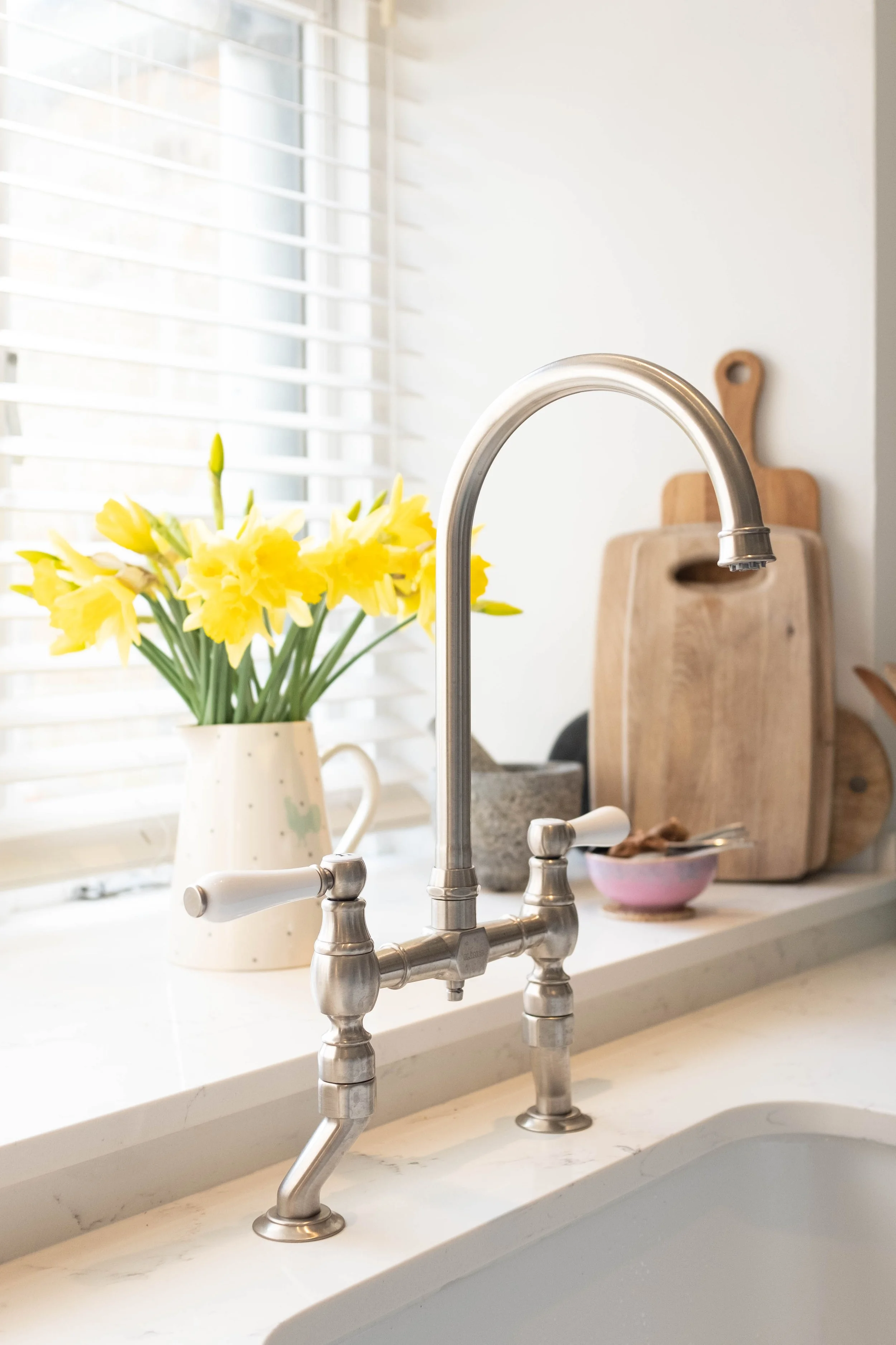 Close-up of a kitchen sink faucet with a vase of yellow daffodils on the window sill behind it, along with a wooden cutting board and a pink bowl on the counter.