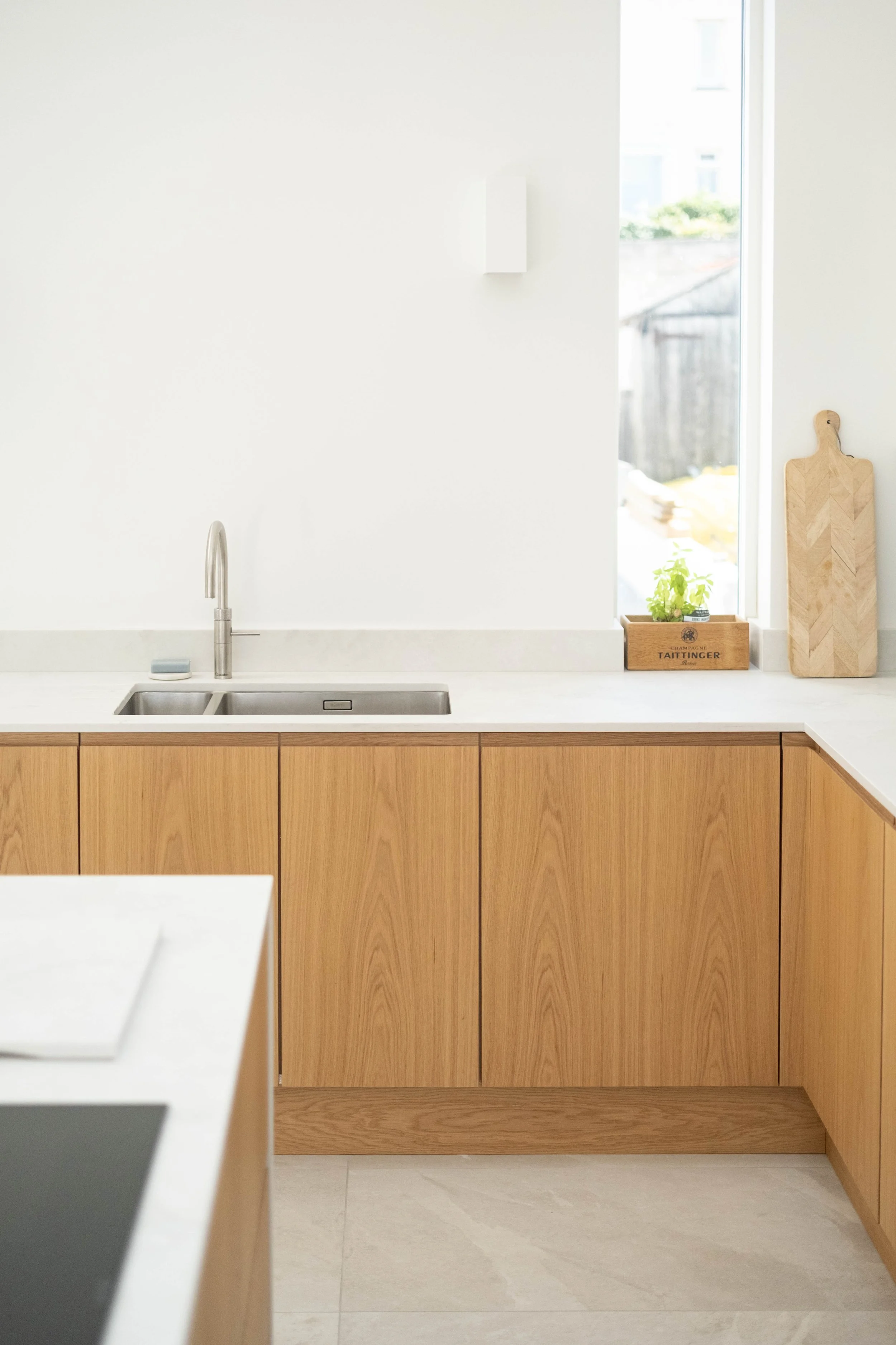 A modern kitchen with a white countertop, wooden cabinets, a stainless steel sink, and a faucet. There's a window with sunlight coming in, and on the counter beside the window, there's a small plant in a wooden box and a decorative wooden cutting boa