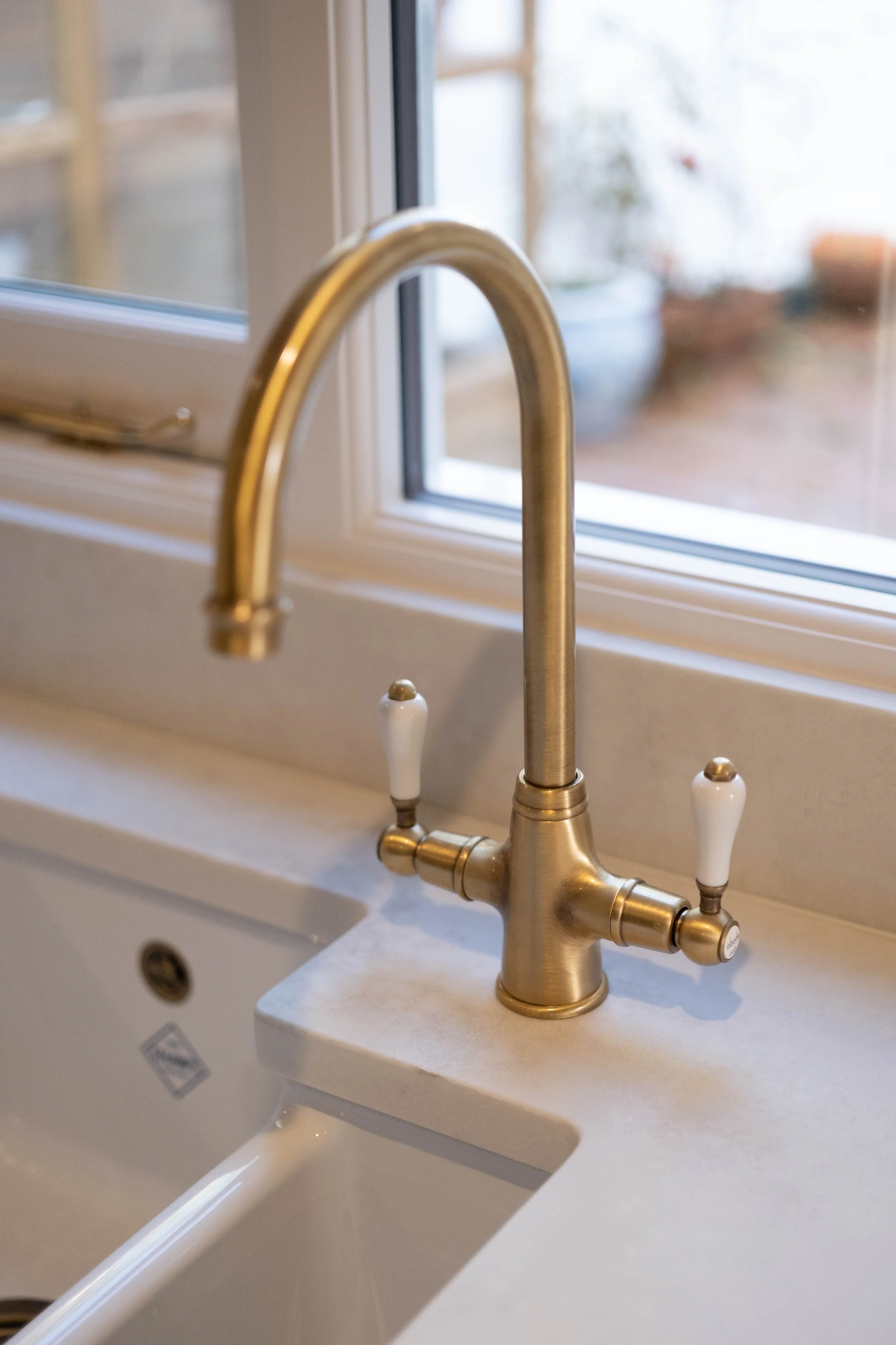 Close-up of a brass kitchen faucet with white handles, mounted on a white sink by a window with a blurred garden view in the background.