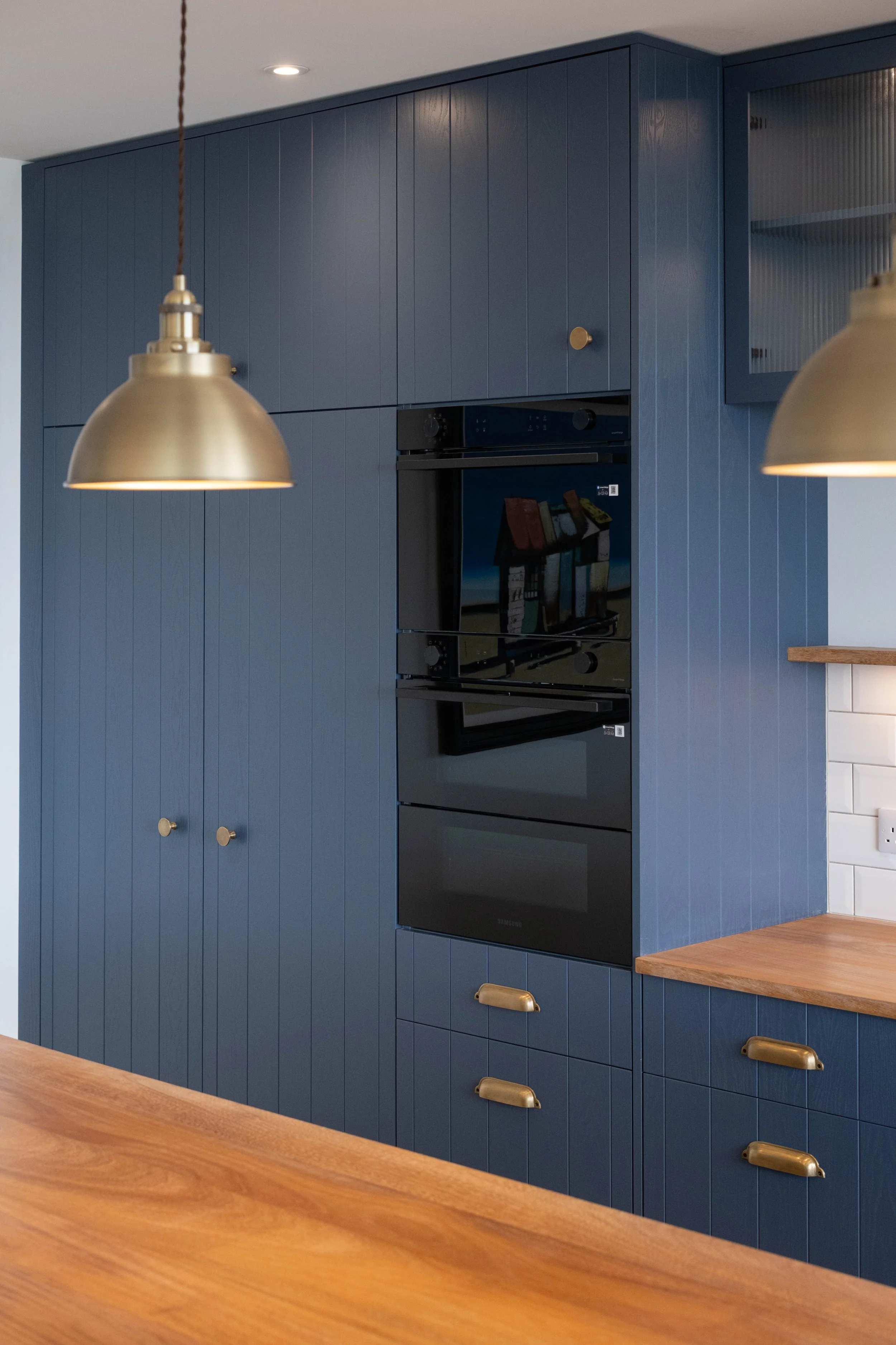 Close-up of a kitchen with blue wooden cabinets, a black oven, and brass hardware, with a wooden countertop in the foreground and two gold pendant lights hanging from the ceiling.