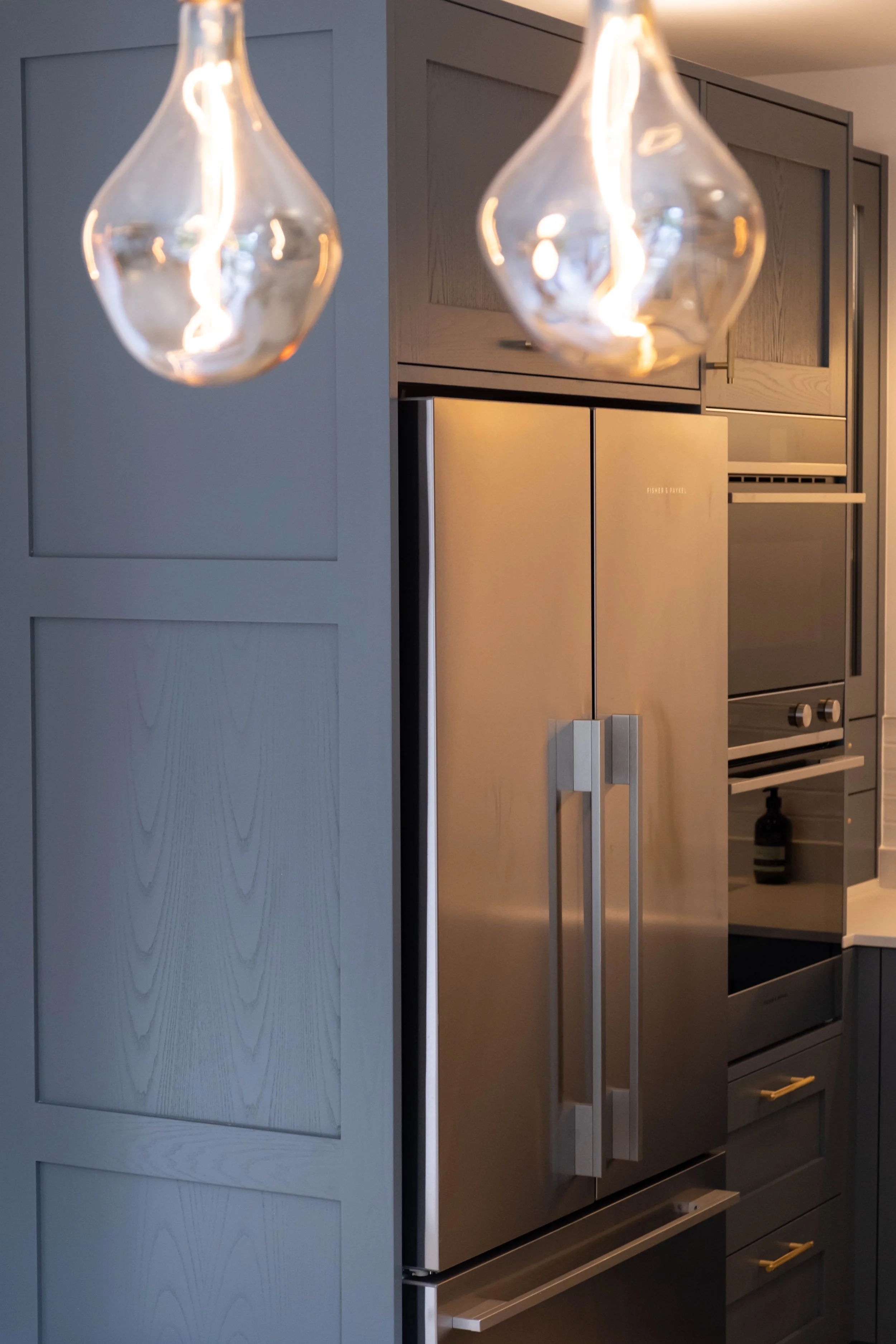 Close-up of two pendant lights with glass shades hanging over a kitchen countertop, with kitchen cabinets and appliances in the background.