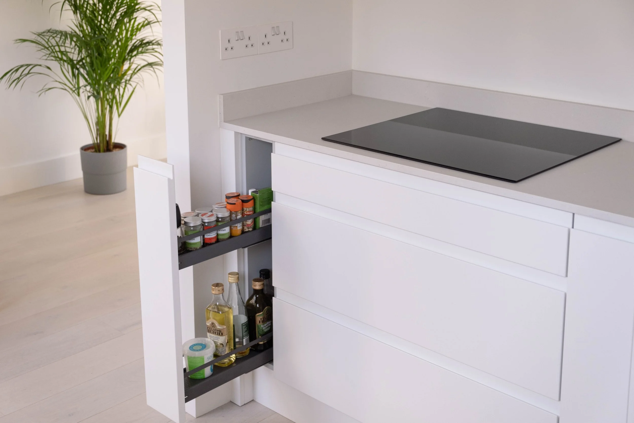 Kitchen corner with white cabinets, black electric cooktop, potted plant, and open spice drawer showing jars and bottles.