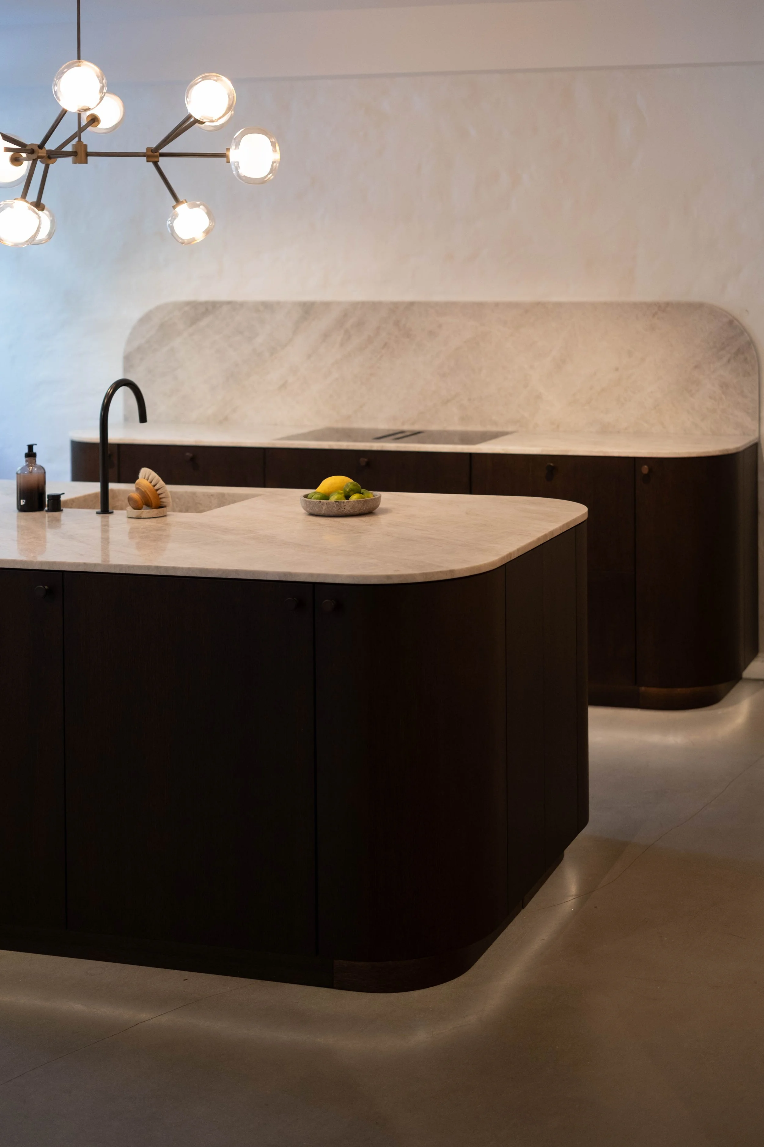 Modern kitchen with dark wood cabinets, a light marble countertop, a sink with a black faucet, and a decorative bowl of lemons and limes. There is a contemporary chandelier with multiple exposed bulbs hanging from the ceiling.